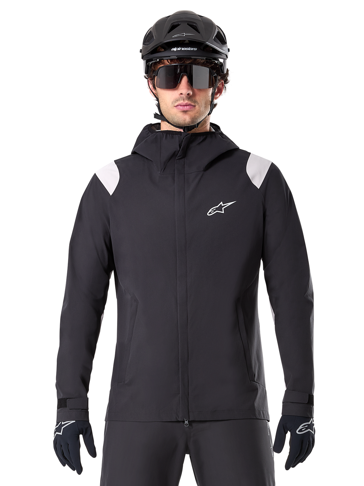 Casaco Impermeável Alpinestars A-Dura, casaco de chuva para MTB, preto com painéis brancos nos ombros, usado com capacete de MTB preto, óculos de sol e luvas, design impermeável com capuz e fecho frontal, equipamento desportivo de exterior para ciclismo