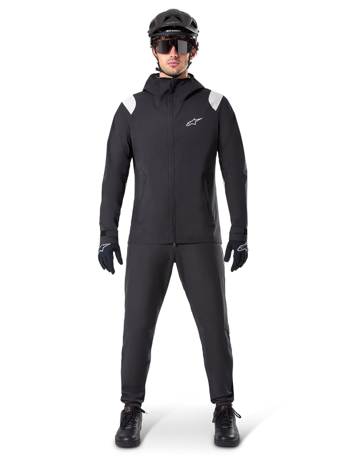 Casaco de Chuva Alpinestars A-Dura, casaco de chuva para mountain bike, preto com detalhes brancos nos ombros e logótipo branco no peito, usado com calças pretas, capacete preto, óculos de sol e luvas, equipamento de mountain bike coordenado para tempo chuvoso