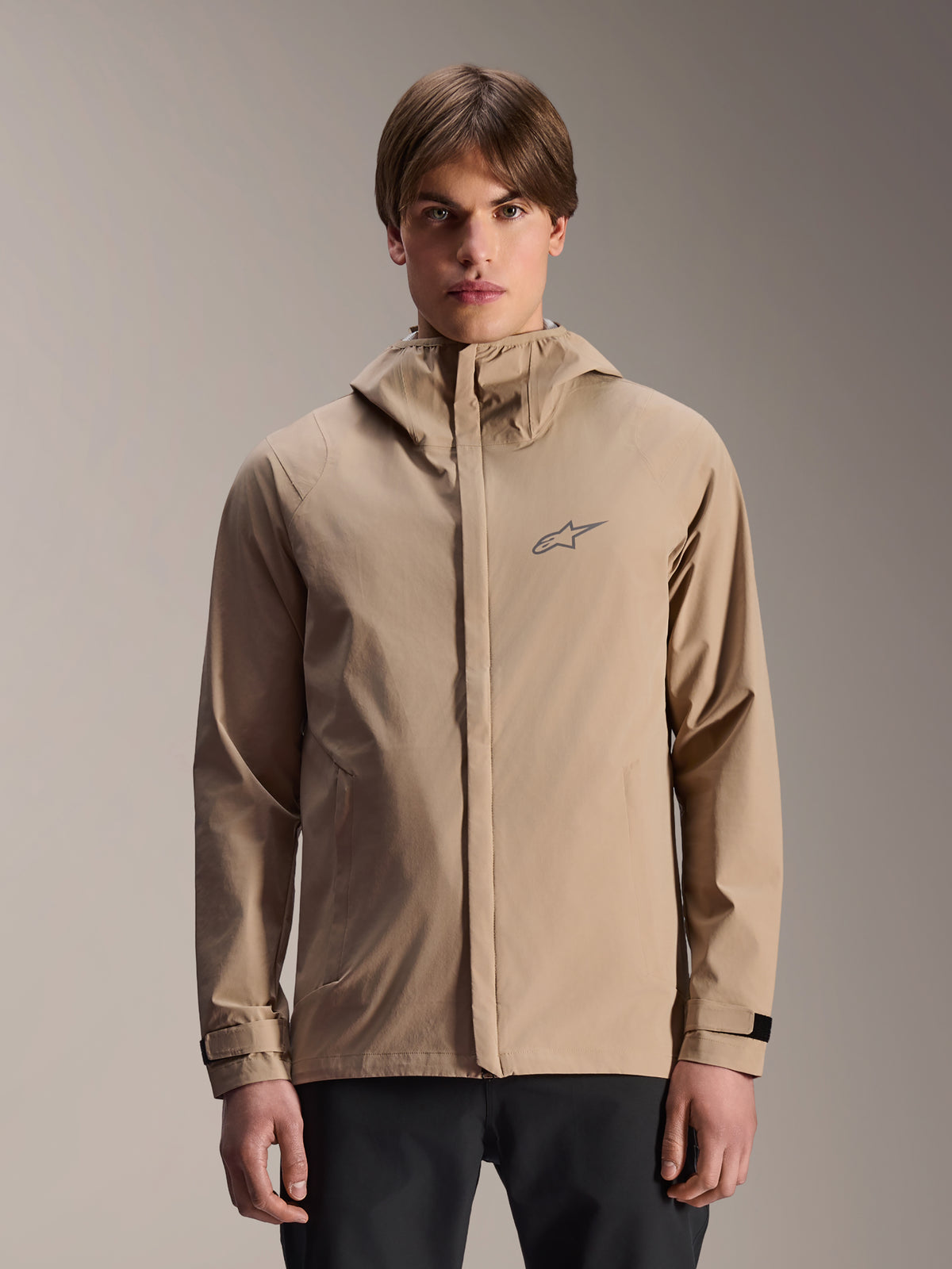 Alpinestars A-Dura Rain Jacket, casaco de btt impermeável com capuz em castanho claro, vestido por um modelo, com um pequeno logótipo no peito, punhos ajustáveis e fecho frontal oculto, equipamento de ciclismo impermeável