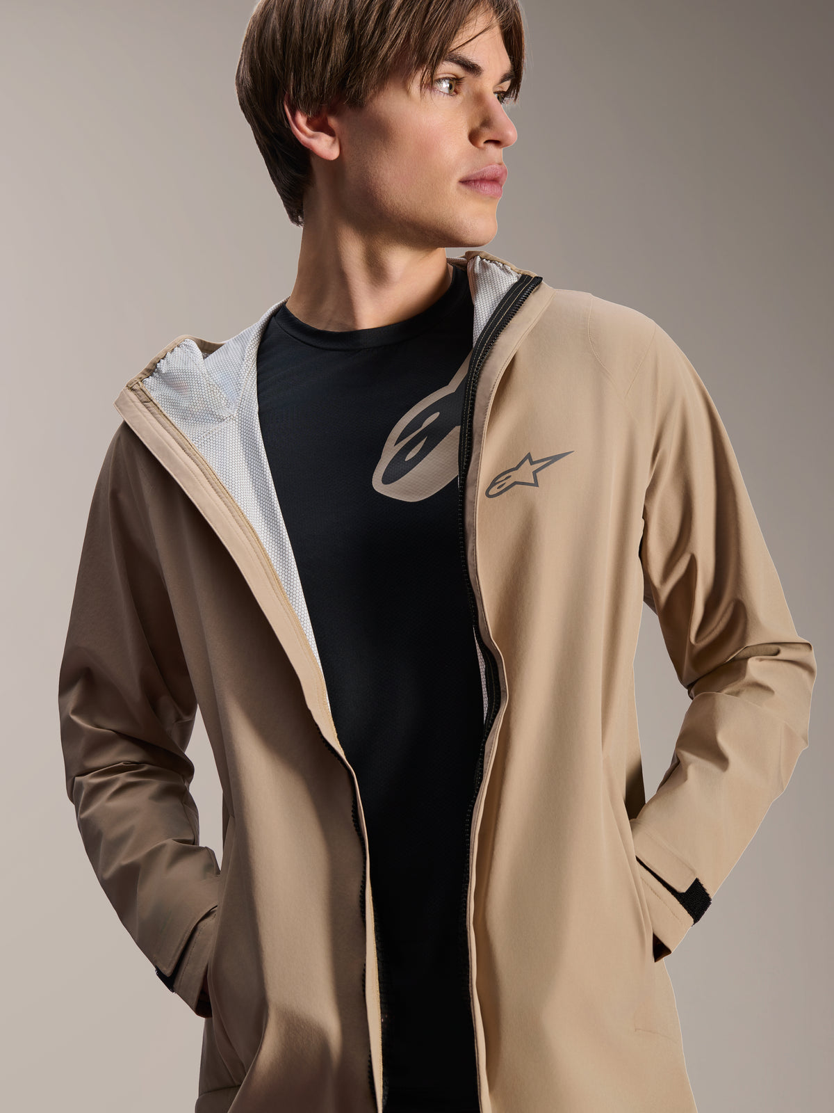 Modelo masculino a usar o Casaco Impermeável Alpinestars A-Dura, castanho claro, aberto sobre uma t-shirt preta, mostrando o forro de malha branco, casaco de ciclismo versátil e resistente às intempéries