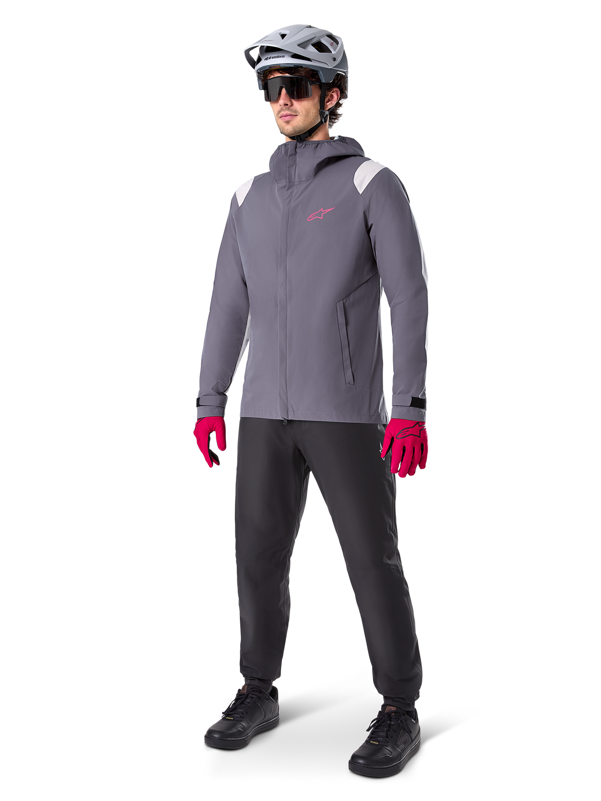 Modelo masculino a usar o Casaco de Chuva Alpinestars A-Dura em Dark Titanium, casaco cinzento com capuz, logótipo cor-de-rosa no peito e painéis cinzento claro nos ombros, combinado com um capacete de MTB cinzento, óculos de sol escuros, luvas cor-de-rosa vibrantes e calças pretas para ciclismo todo-o-terreno