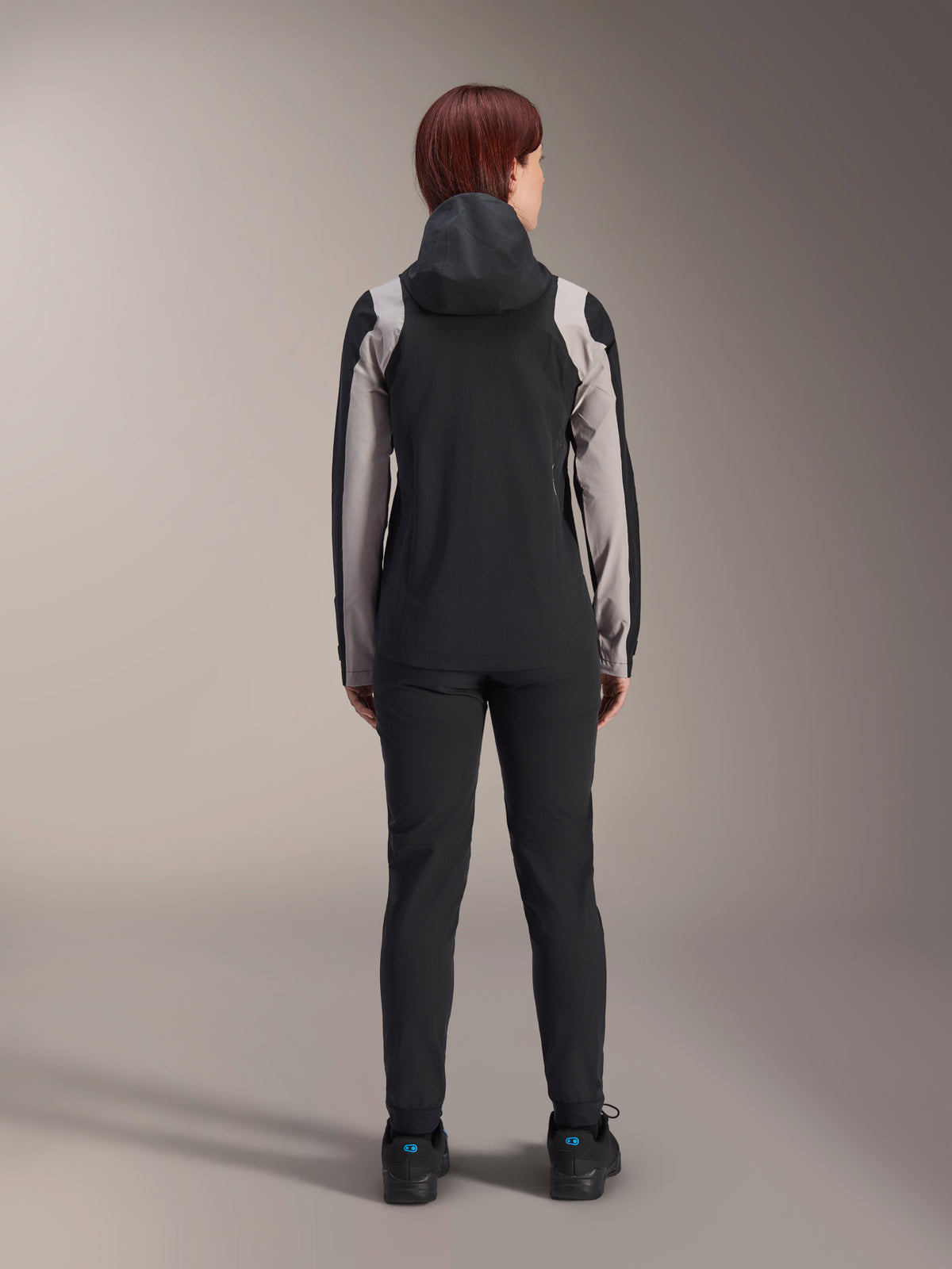 Female rider from behind wearing Alpinestars Stella A-Dura Rain Jacket, Black with light grey panels on the sleeves and shoulders, hood up, worn with black pants and cycling shoes, technical mountain bike rain gear