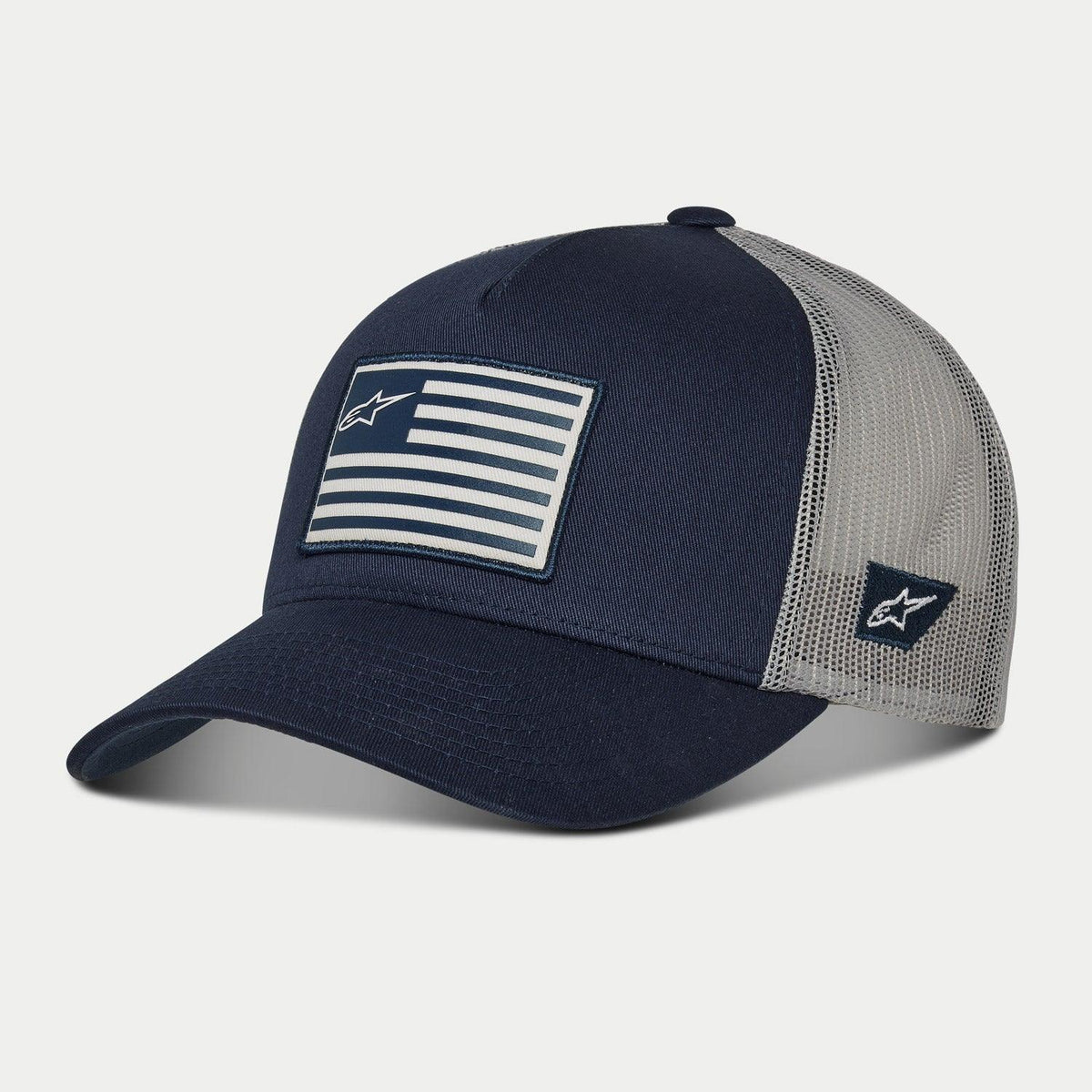 Boné Alpinestars Flag Snapback, azul marinho e cinzento, pala curva com copa estruturada, patch de bandeira impresso na frente e logótipo bordado na lateral, parte traseira em malha para respirabilidade, estilo casual inspirado no desporto motorizado