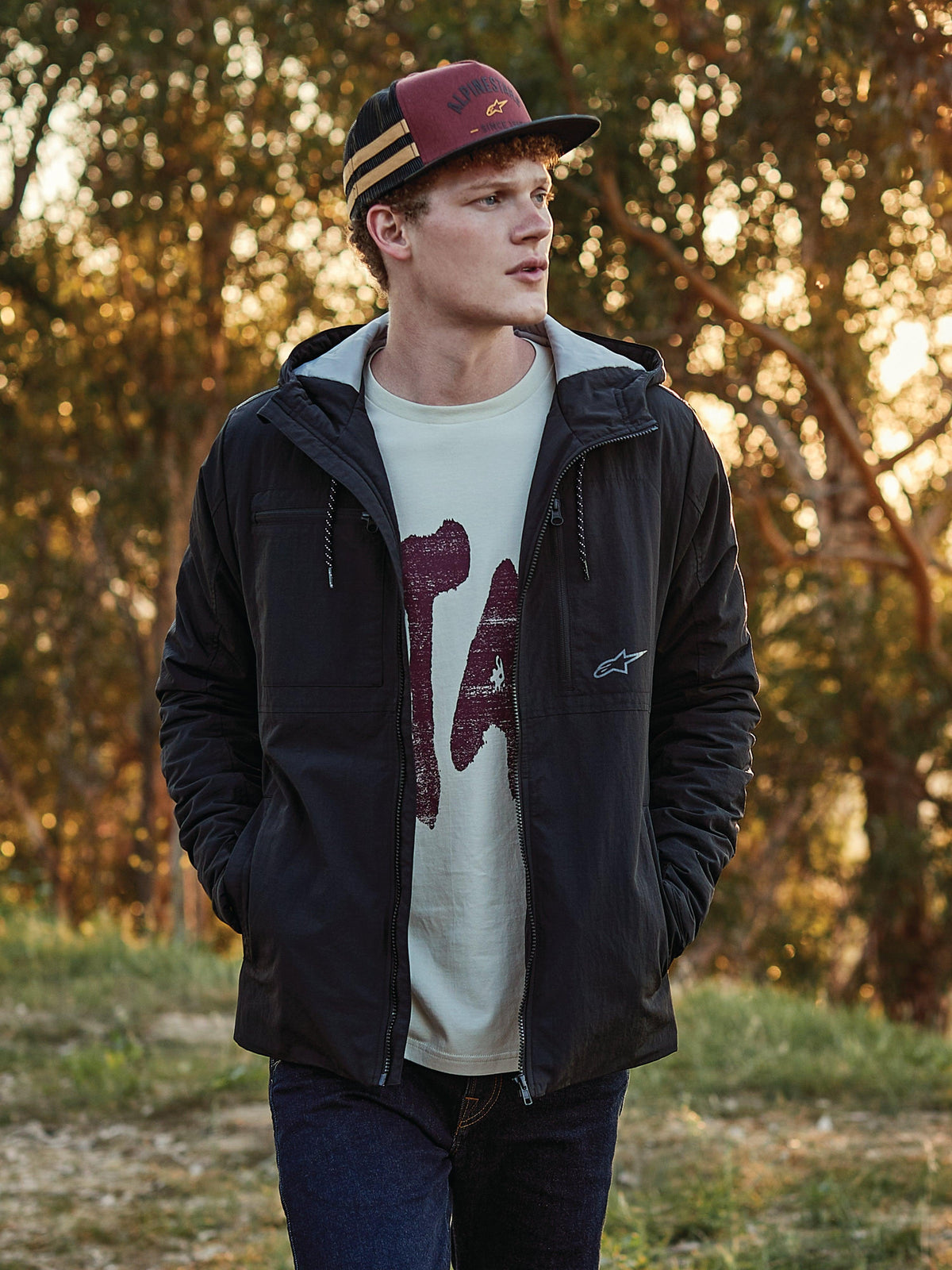 Alpinestars Backline Trucker Hat in Brick, Black, and Mustard, worn by a man paired with a black hooded jacket and graphic t-shirt, casual motorsport-inspired lifestyle streetwear in an outdoor setting at golden hour.