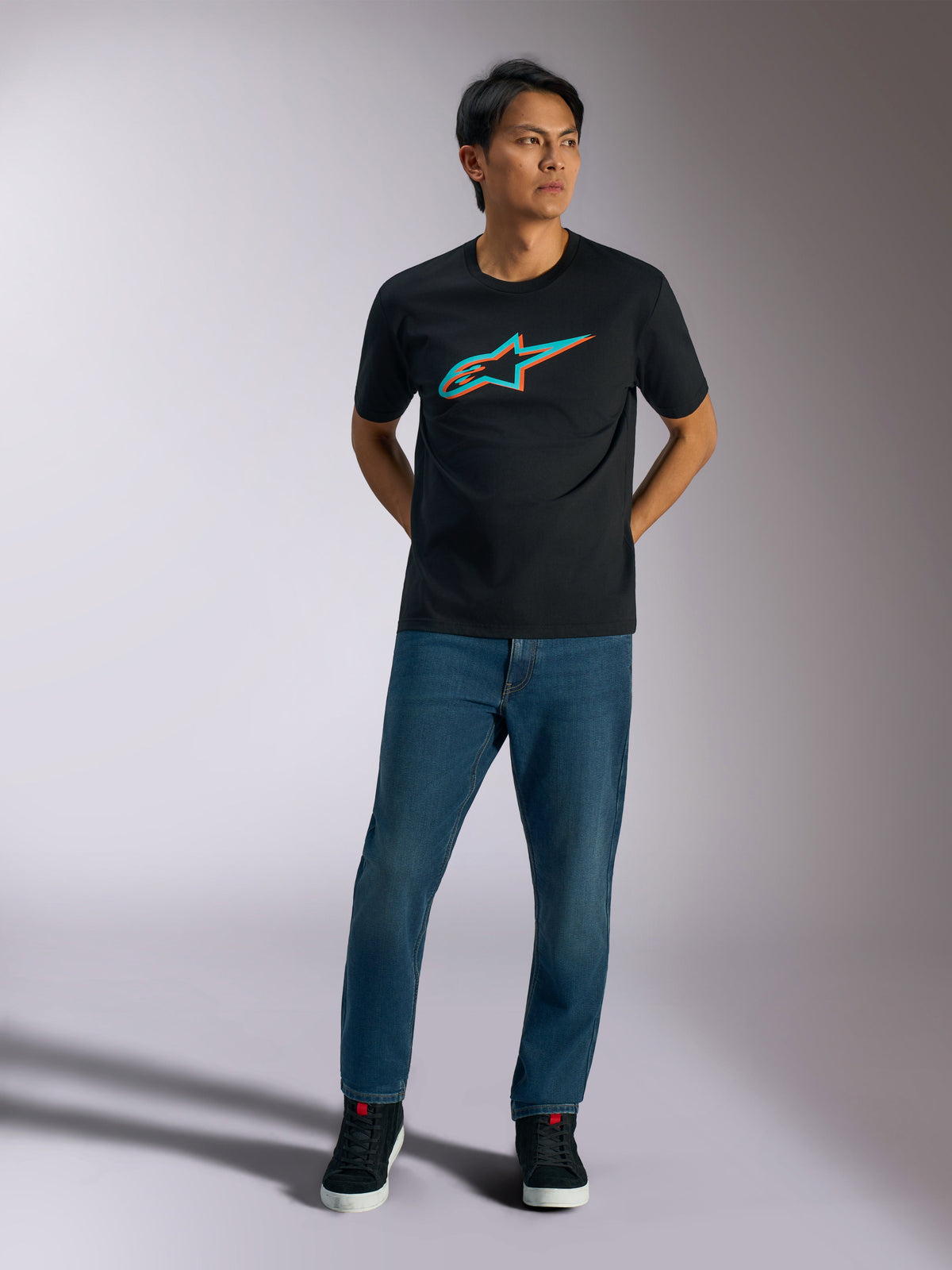 T-shirt Alpinestars Ageless Shadow CSF, t-shirt de manga curta, preta, logótipo Ageless shadow em turquesa e laranja no peito, vestida por um modelo com calças de ganga azuis e sapatos pretos, estilo casual motorsport