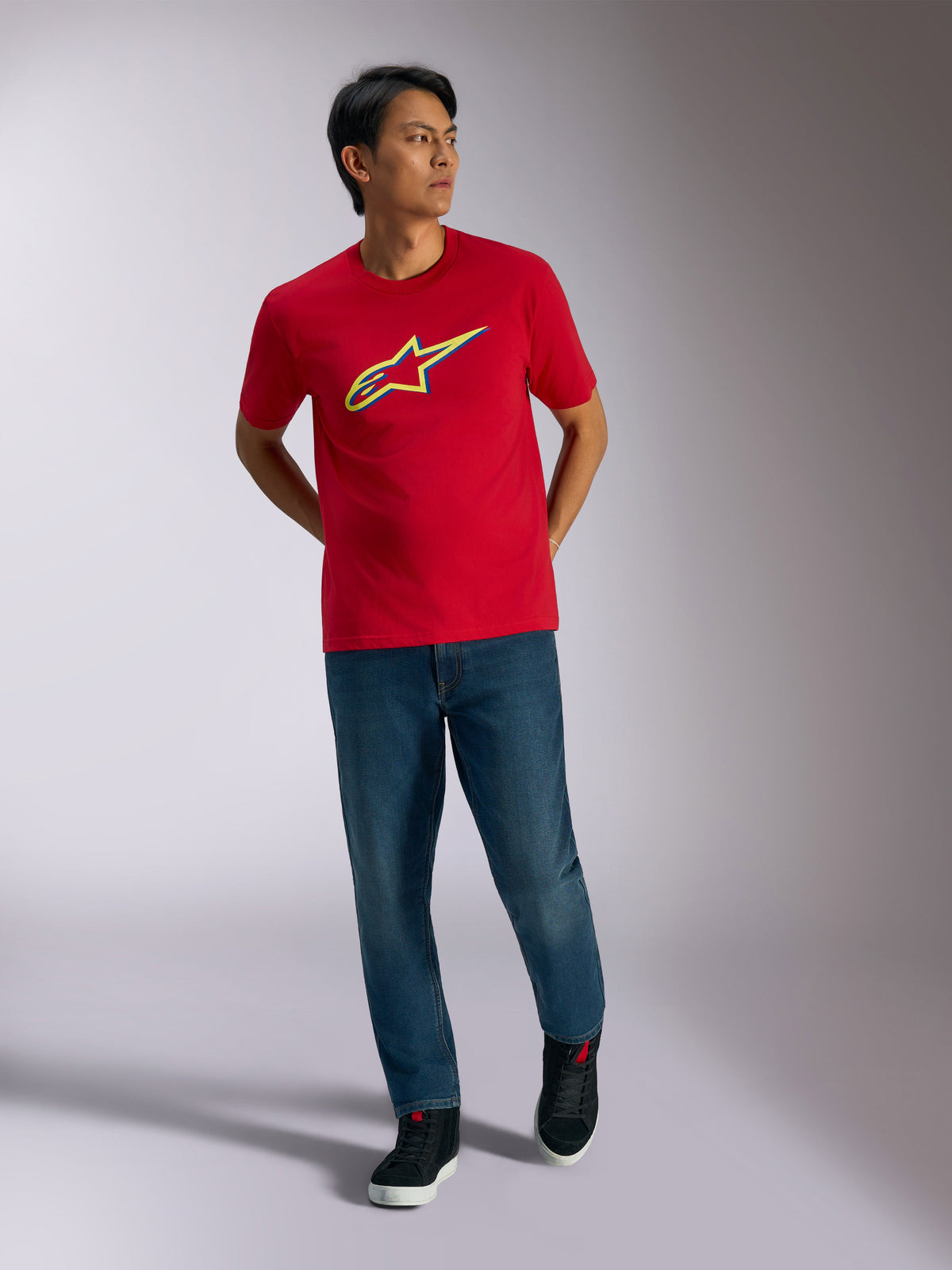 Alpinestars Ageless Shadow CSF Tee, t-shirt vermelha de manga curta, logótipo gráfico Ageless shadow em amarelo e azul, usada com calças de ganga azuis e sapatilhas pretas, vestuário casual de estilo de vida motorsport
