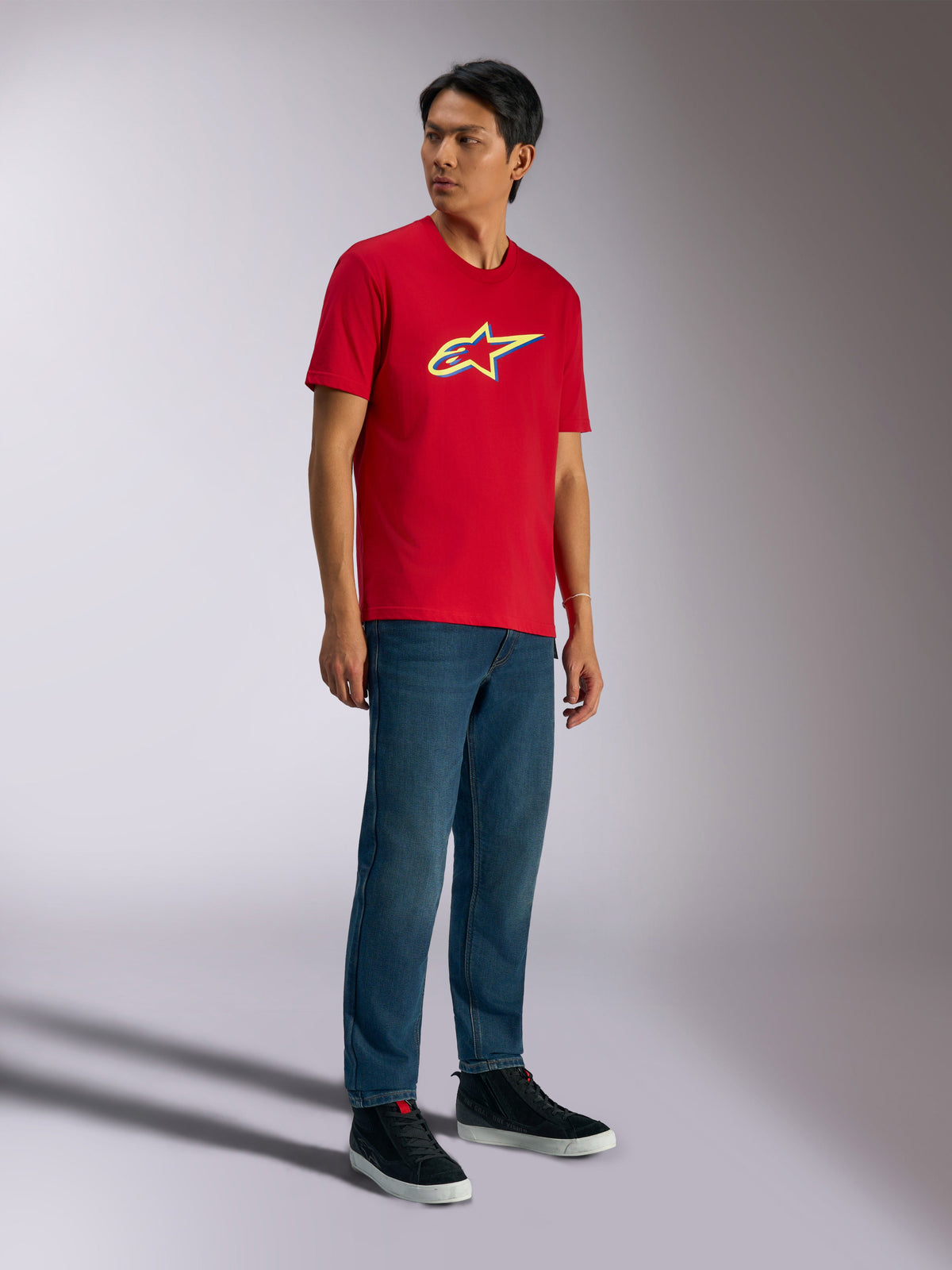 Alpinestars Ageless Shadow CSF Tee, t-shirt de manga curta, vermelha, com logótipo Ageless shadow com detalhes em amarelo e azul, usada por modelo com calças de ganga e sapatilhas pretas, casual motorsport streetwear