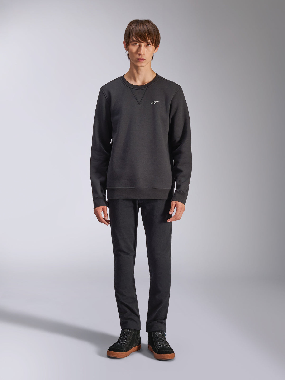 Sweatshirt Alpinestars Positive Crew, gola redonda casual preta, pequeno logótipo branco no peito, detalhe de costura em V na gola, fotografia de corpo inteiro em estúdio com modelo a usar calças pretas e sapatilhas, vestuário de estilo de vida inspirado no desporto motorizado