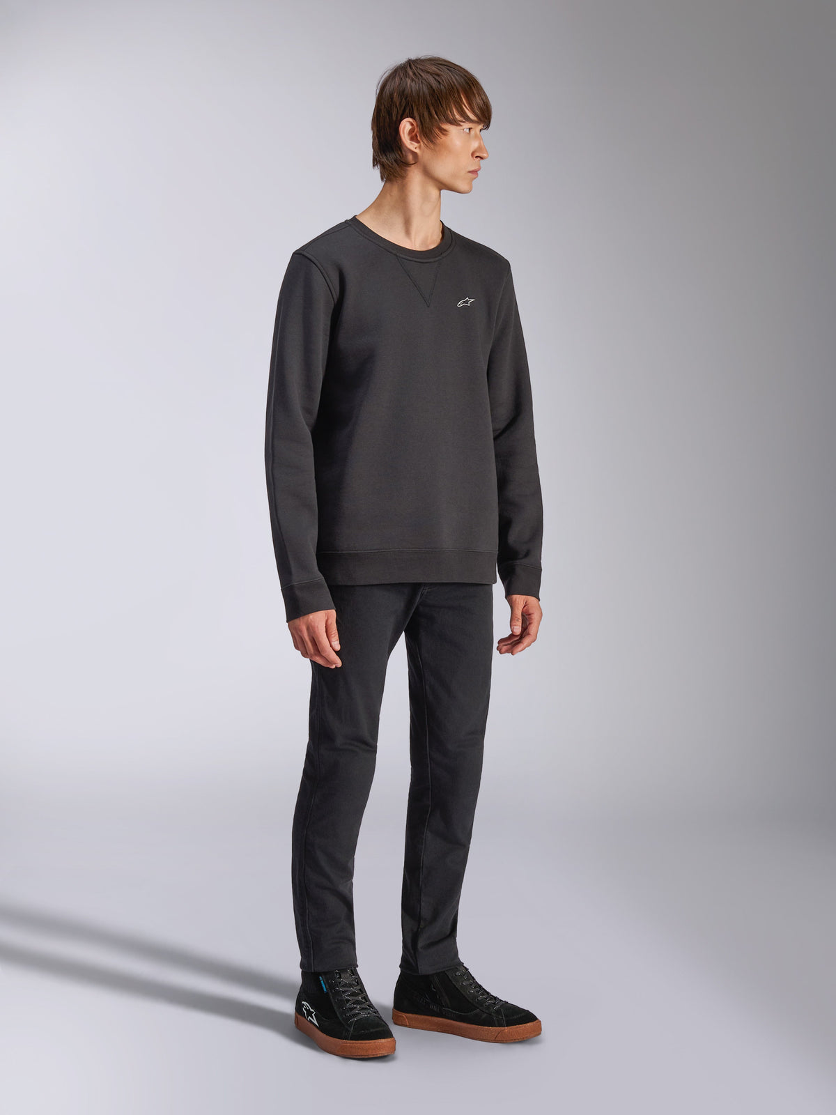 Alpinestars Positive Crew, sweatshirt preta, pequeno logótipo Alpinestars branco no peito, gola canelada com detalhe de costura em V, punhos e bainha canelados, usada por um modelo com calças pretas e ténis, streetwear casual inspirado no desporto motorizado