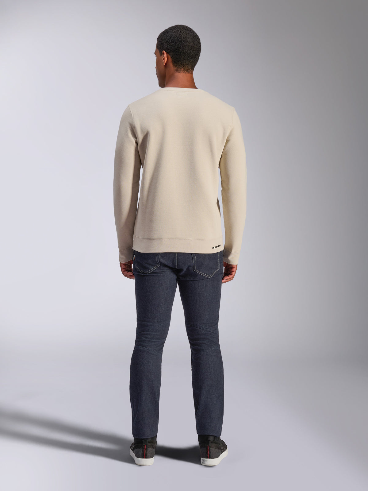 Sweatshirt Alpinestars Positive Crew, Off White, vista traseira de um homem a usar uma camisola bege de gola redonda com bainha e punhos canelados, combinada com calças de ganga escuras, streetwear casual inspirado no desporto motorizado.