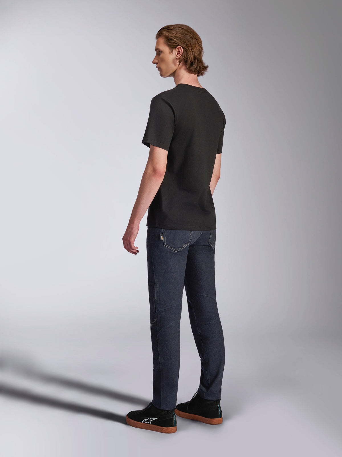 Alpinestars Luxo CSF Tee, t-shirt de manga curta, preta, vista traseira no modelo combinada com calças de ganga azul-escuro e sapatilhas pretas, streetwear casual inspirado no desporto motorizado
