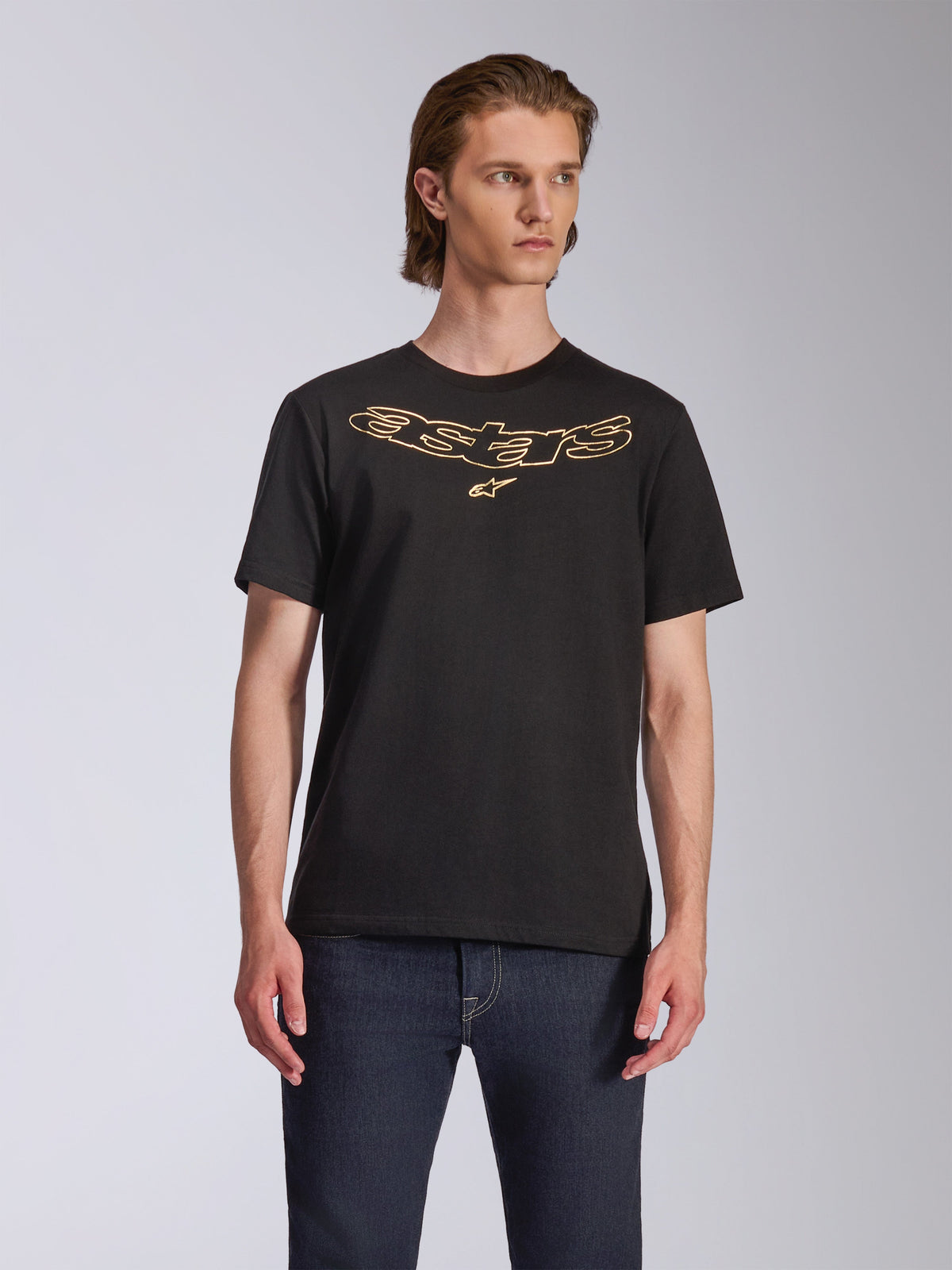 T-shirt Alpinestars Luxo CSF, t-shirt de manga curta, preta, logótipo Astars com contorno dourado no peito, usada com calças de ganga azul-escuro, streetwear casual de desportos motorizados