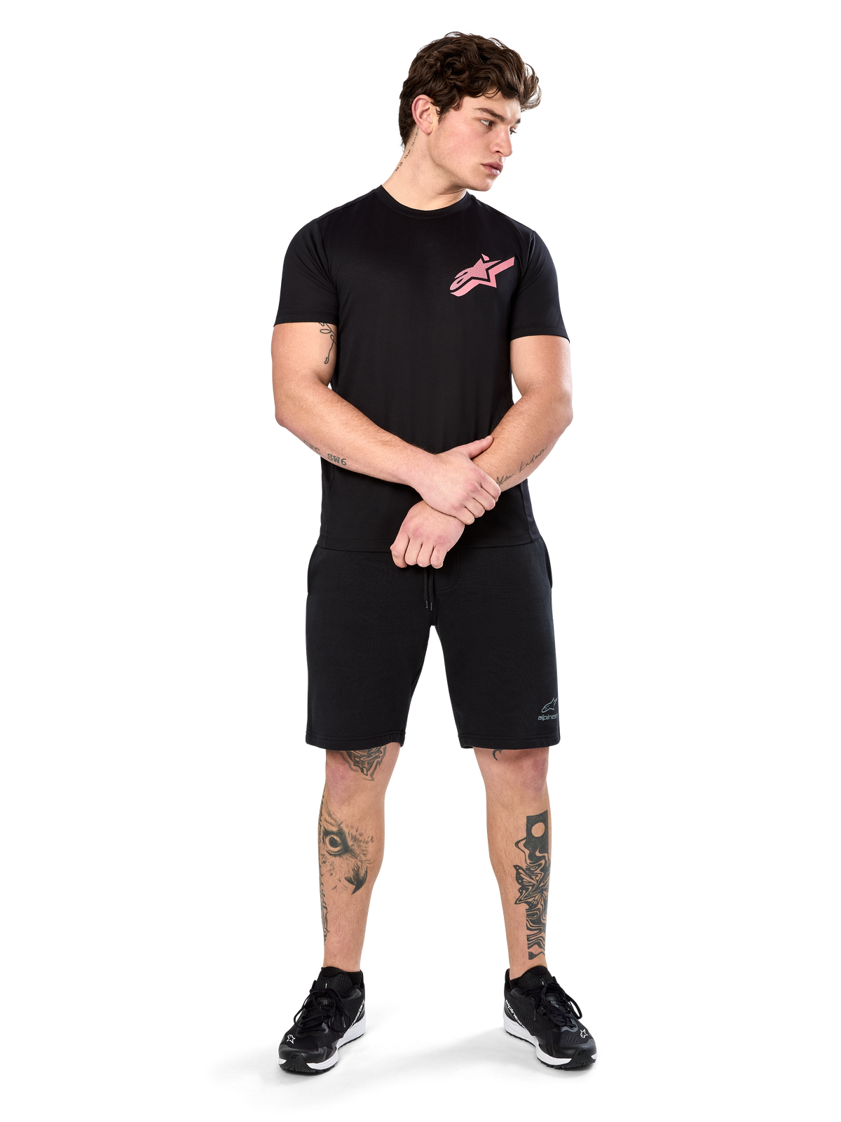 Modelo a usar a t-shirt Alpinestars Attrition Performance, t-shirt de performance de manga curta, preta com logótipo Astars cor-de-rosa no peito, combinada com calções pretos e ténis, roupa desportiva casual inspirada no desporto motorizado