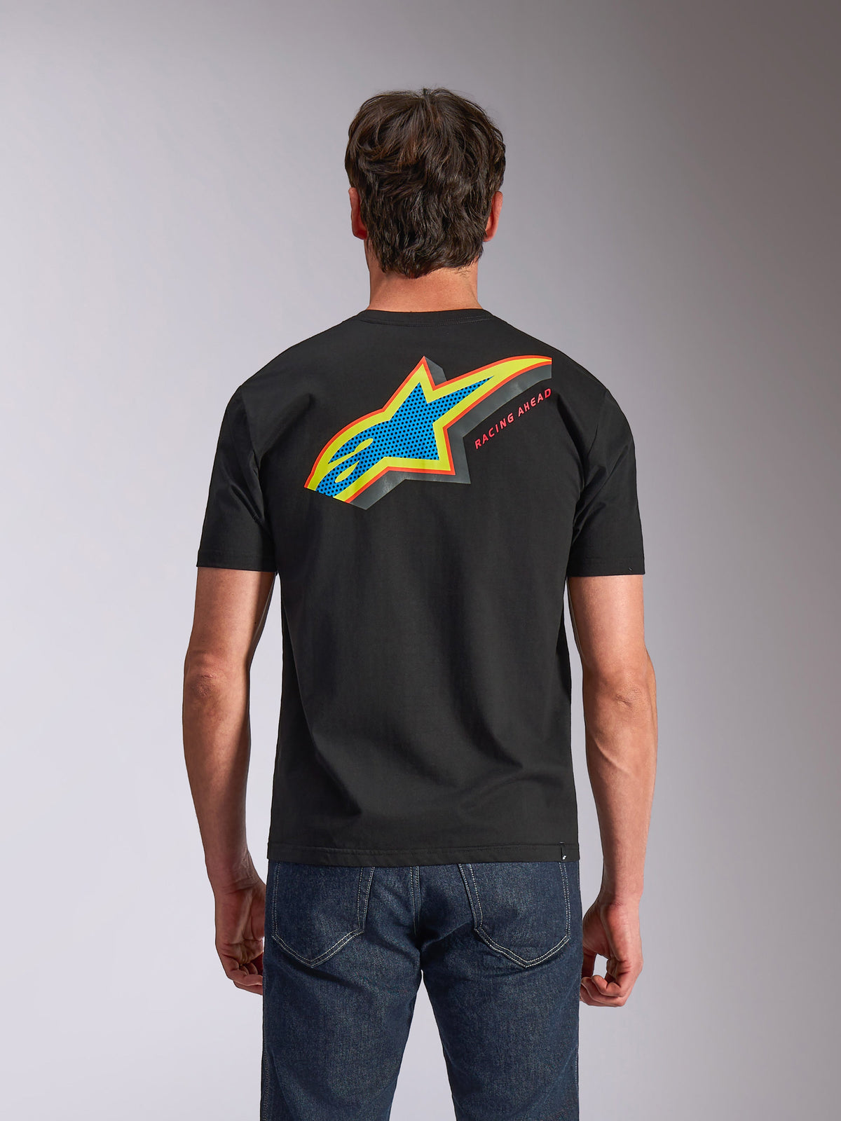 T-shirt Alpinestars Angler CSF, de manga curta, preta, vista de trás mostrando o logótipo Astars vibrante e multicolor com preenchimento perfurado e o texto 'RACING AHEAD', usada com calças de ganga azuis, vestuário casual inspirado no desporto motorizado
