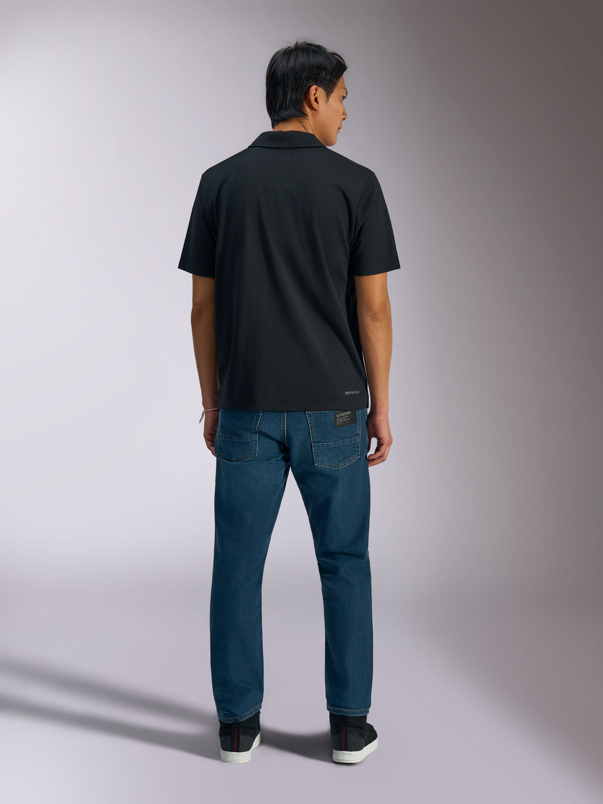 Alpinestars Polo Impetus Sport, preto, vista traseira de polo de manga curta com a marca Alpinestars na parte inferior das costas, usado com calças de ganga azuis, vestuário desportivo casual inspirado no desporto motorizado