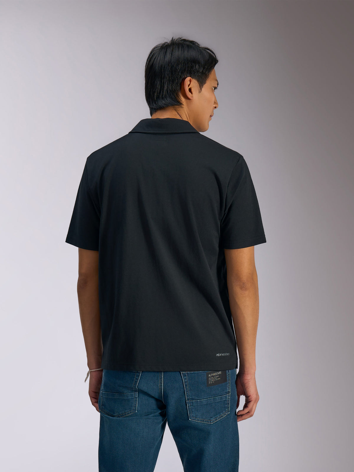 Alpinestars Impetus Sport Polo, polo de manga curta preto, vista traseira de homem a usar o polo com calças de ganga azuis, com pequena marca nominativa Alpinestars na parte inferior das costas, vestuário desportivo casual inspirado no desporto motorizado