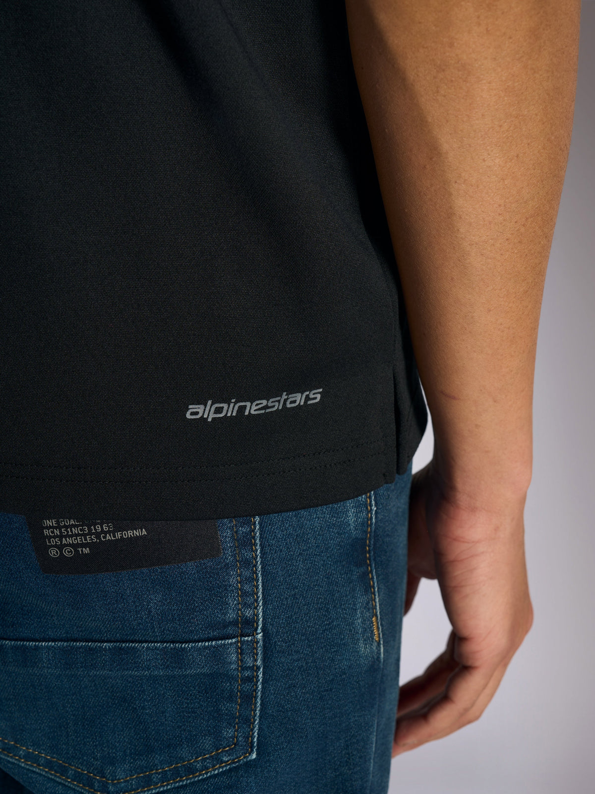 Polo Alpinestars Impetus Sport, vista detalhada da bainha inferior e abertura lateral, preto com logótipo Alpinestars a cinzento, vestuário desportivo casual inspirado no desporto motorizado