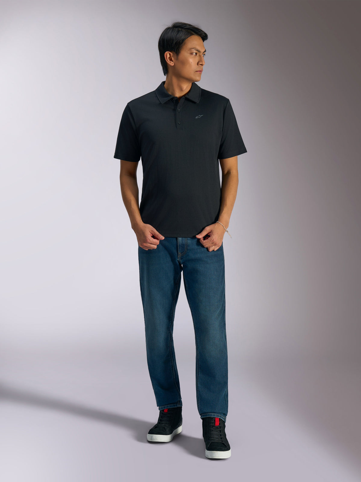 Alpinestars Impetus Sport Polo, polo de manga curta preto, apresenta uma carcela de três botões e o logótipo Astars cinzento no peito, usado por um homem com calças de ganga azuis e sapatilhas de cano alto pretas, roupa desportiva casual inspirada no desporto motorizado