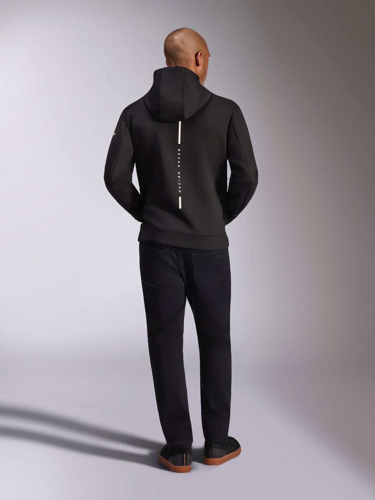 Alpinestars Insight Hoodie, preto, vista traseira no modelo, com um logótipo de risca vertical branca nas costas com o texto "Racing Ahead", sweatshirt casual com capuz em lã tri-blend de dupla face, combinada com calças pretas.