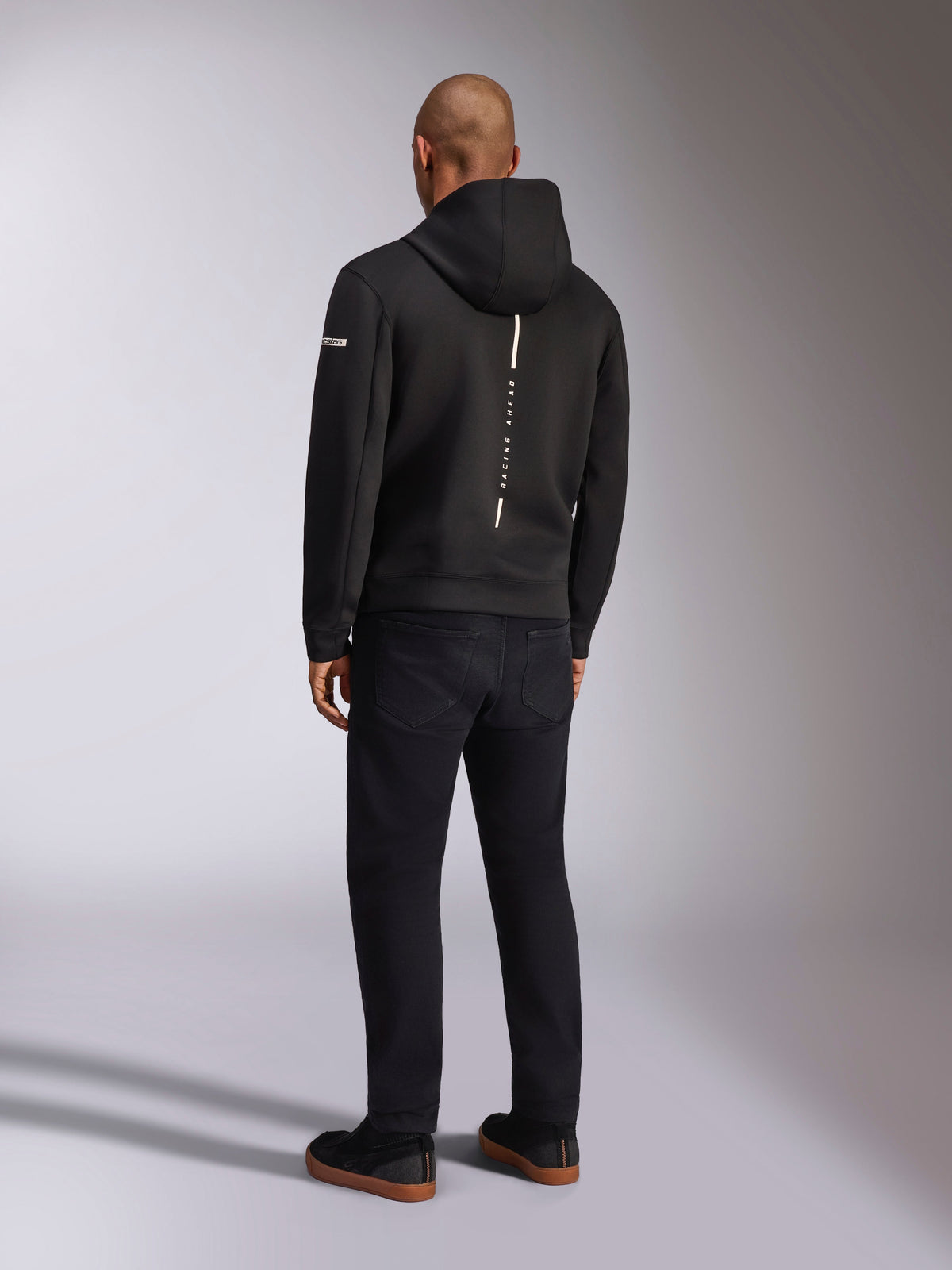 Alpinestars Insight Hoodie, sweatshirt com capuz desportiva preta, vista traseira mostrando uma risca vertical branca com a marca 'RACING AHEAD', logótipo na manga, combinada com calças pretas e ténis, design casual inspirado no desporto motorizado