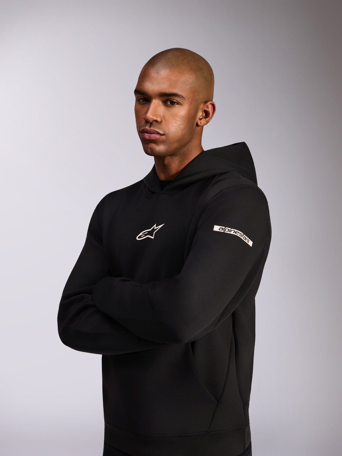 Alpinestars Insight Hoodie, sweatshirt casual de sportswear, preto, logótipo Astars branco no peito e logótipo escrito na parte superior da manga, modelo a usar o hoodie com os braços cruzados, confeção premium em lã de dupla face