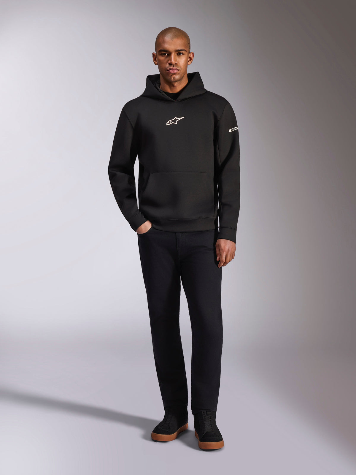Alpinestars Insight Hoodie, sweatshirt com capuz preta com logótipo Astars branco no peito e na manga, vestida por um modelo com calças pretas e sapatilhas pretas, streetwear casual inspirado no desporto motorizado