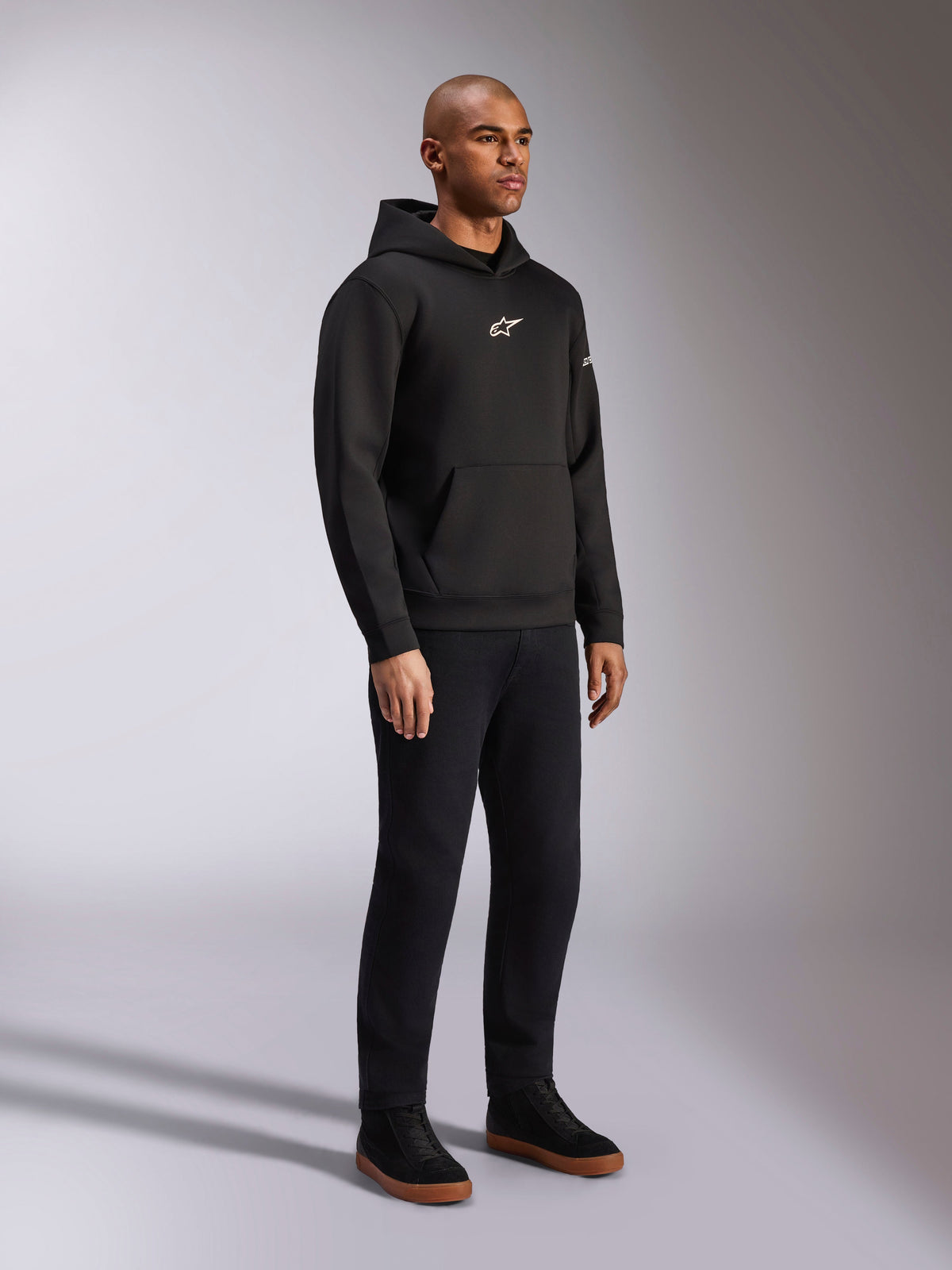 Alpinestars Insight Hoodie, preto, sweatshirt casual de lã com capuz e logótipo Astars branco no peito, bolso canguru, usado com calças pretas e sapatilhas, fotografia de estúdio de corpo inteiro, streetwear inspirado no desporto motorizado