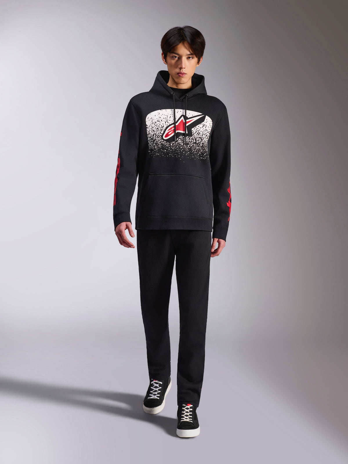 Camisola com capuz Alpinestars Obsessed, sweatshirt preta, logótipo Astars grande em branco e vermelho com gradação MX, logótipos vermelhos nas mangas, usada por um modelo num conjunto casual totalmente preto, vestuário de lifestyle inspirado no desporto motorizado