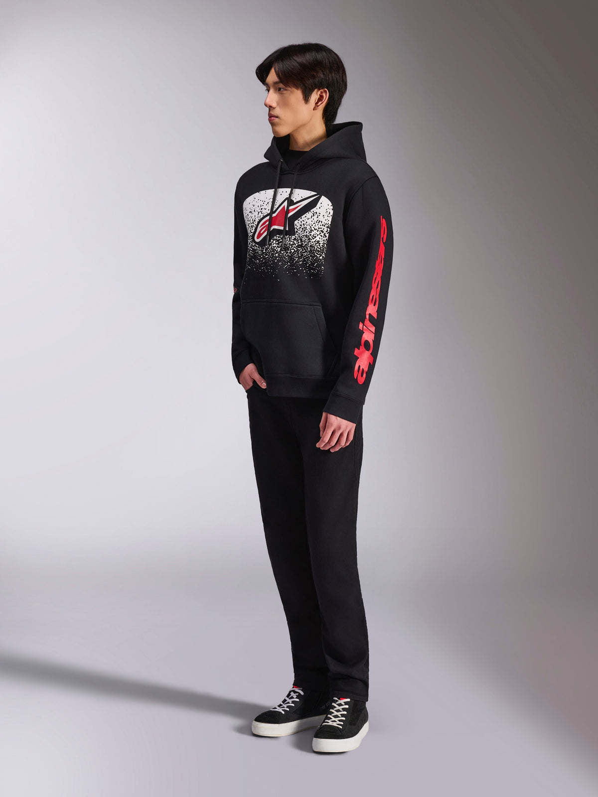 Alpinestars Obsessed Hoodie, camisola com capuz em fleece preto, com um grande grafismo branco salpicado no peito, logótipo Astars vermelho e marca vermelha nas mangas, usada por um modelo com calças pretas e sapatilhas, streetwear casual de desportos motorizados