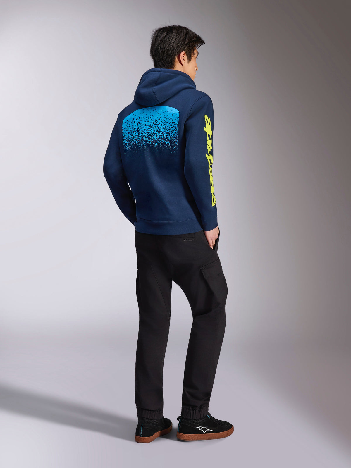 Alpinestars Obsessed Hoodie, sweatshirt casual azul-marinho, vista traseira com gráfico de gradação MX em azul claro e logótipo Alpinestars amarelo na manga, usada com calças pretas e ténis