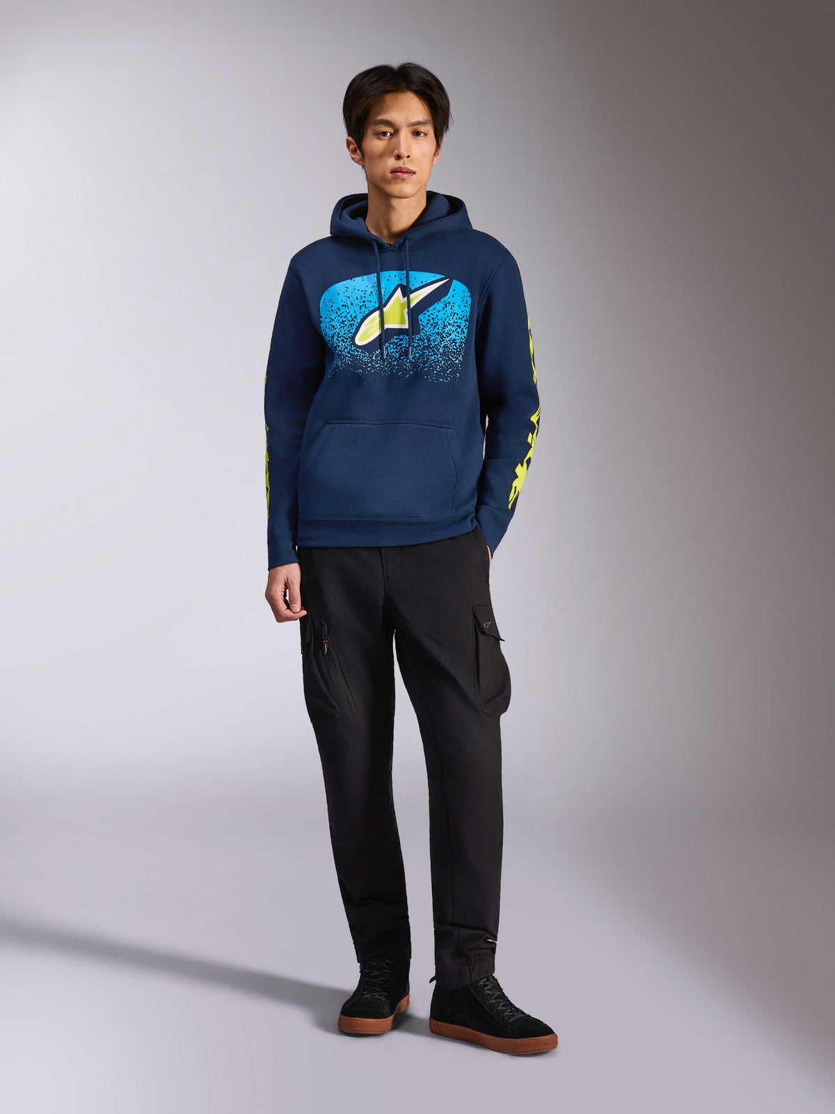 Alpinestars Obsessed Hoodie, camisola com capuz azul-marinho, logótipo Astars frontal com gradação MX azul e estrela amarelo-lima, logótipos Alpinestars em texto amarelo-lima nas mangas, usada por um modelo com calças cargo pretas e ténis pretos, streetwear casual de inspiração motorsport