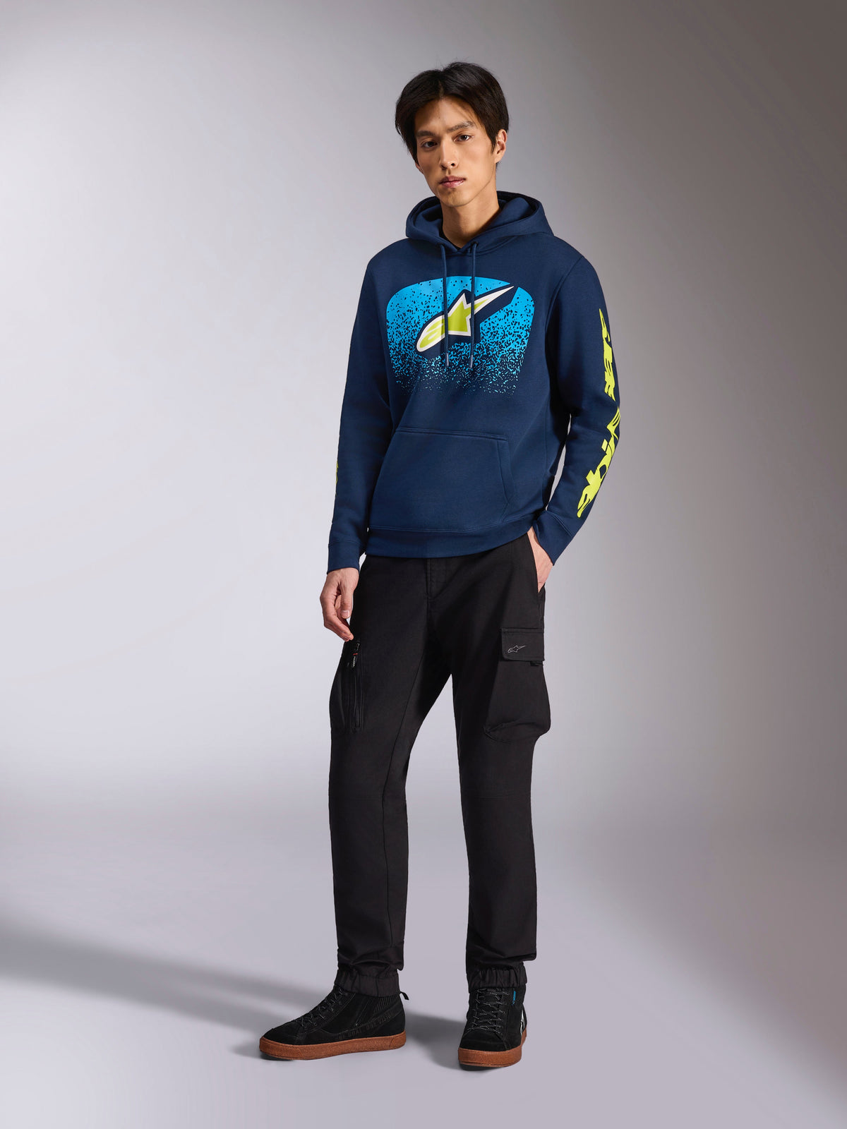 Alpinestars Obsessed Hoodie em azul-marinho, camisola com capuz casual em fleece com grafismo frontal azul-claro e amarelo-lima, logótipo Alpinestars em amarelo-lima na manga, vestida por um modelo com calças cargo pretas e sapatilhas pretas, streetwear de inspiração motorsport