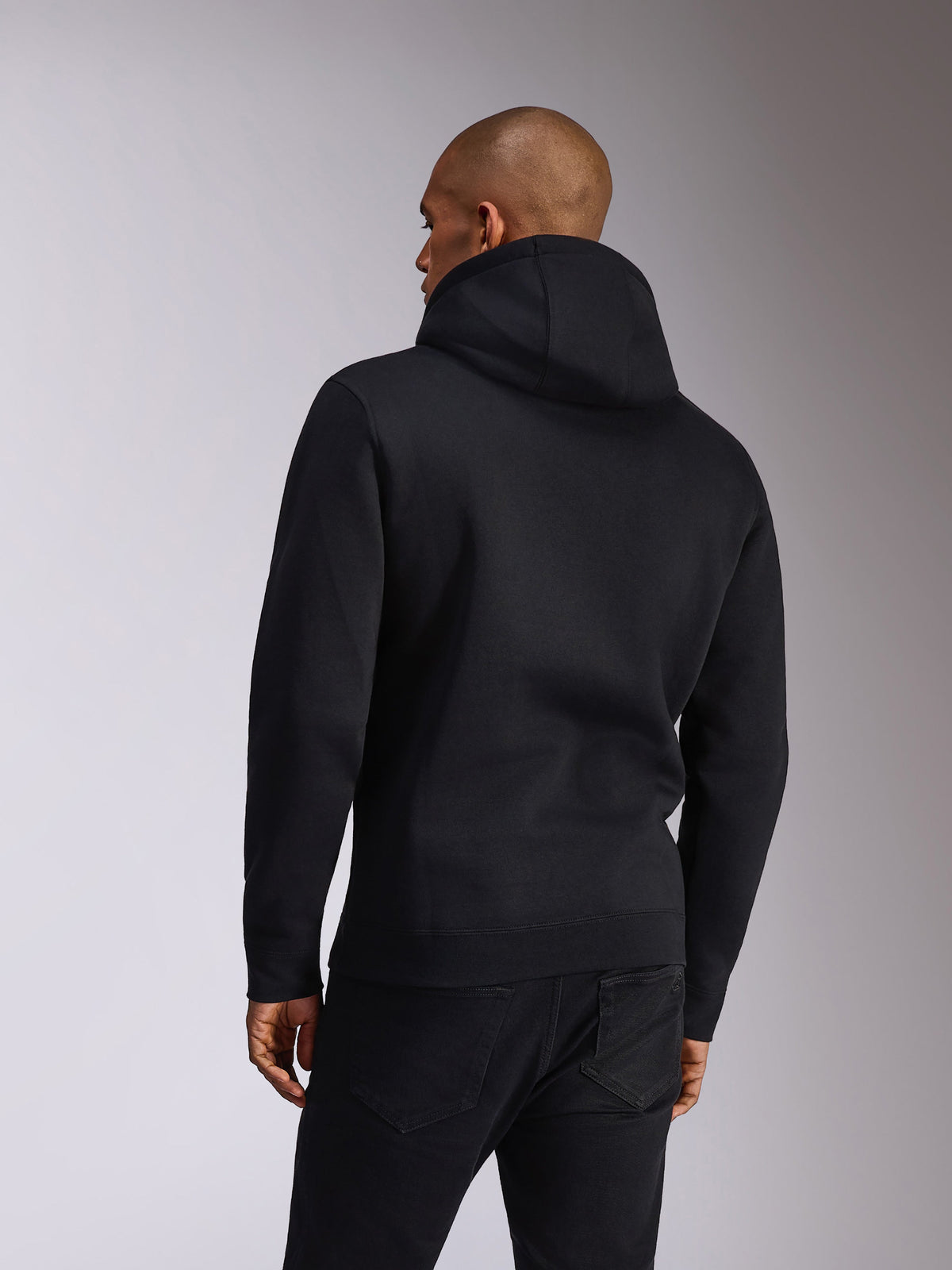 Alpinestars Ellipsoid Hoodie, preto, vista traseira de uma pessoa a usar uma camisola com capuz em fleece, mangas compridas com punhos e bainha canelados, combinada com calças de ganga pretas, vestuário desportivo casual inspirado no desporto motorizado