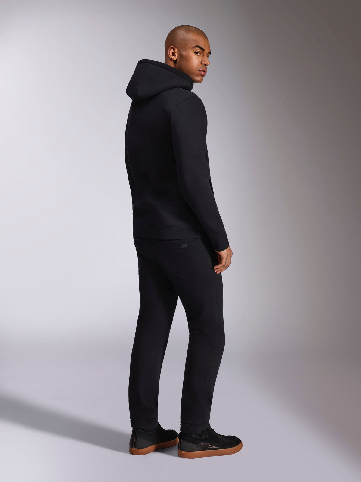 Alpinestars Ellipsoid Hoodie, streetwear casual preto, vista traseira num modelo masculino, camisola com capuz em fleece com punhos e bainha canelados, usada com calças pretas e sapatilhas, design moderno inspirado no desporto motorizado
