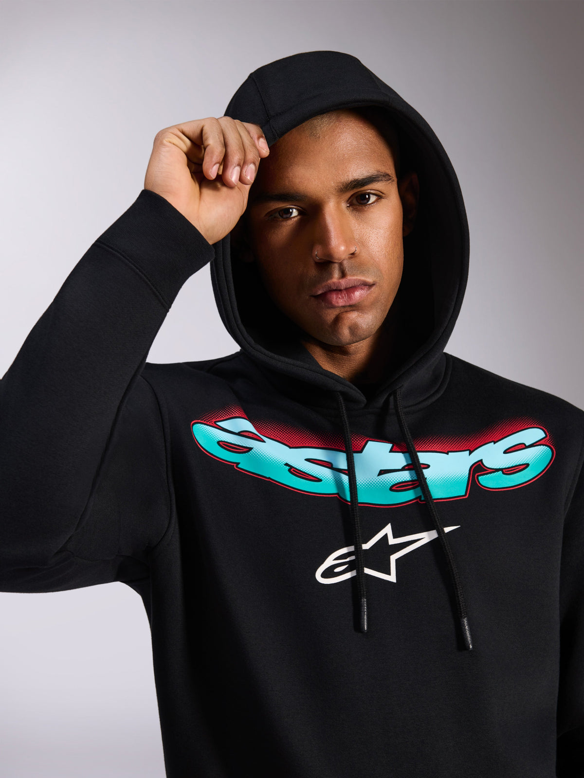 Alpinestars Ellipsoid Hoodie, sweatshirt com capuz em fleece preto, com a palavra "Astars" estilizada num gradiente de azul-turquesa e vermelho e logótipo branco no peito, vestida por um modelo com o capuz levantado, streetwear casual inspirado no desporto motorizado