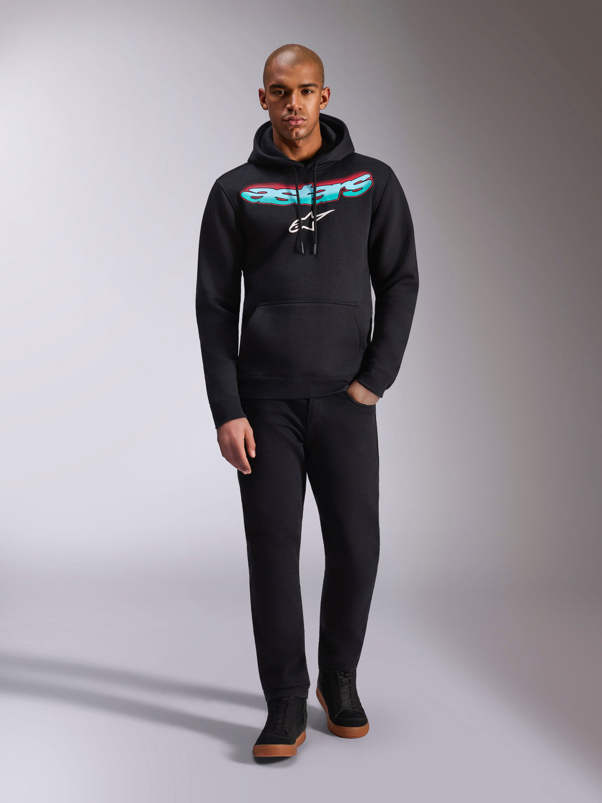 Alpinestars Ellipsoid Hoodie, preta, camisola com capuz em fleece com o logótipo Astars curvo num gradiente de vermelho e azul-petróleo e logótipo da estrela branco, usada por um modelo com calças pretas e sapatilhas, estilo casual motorsport streetwear