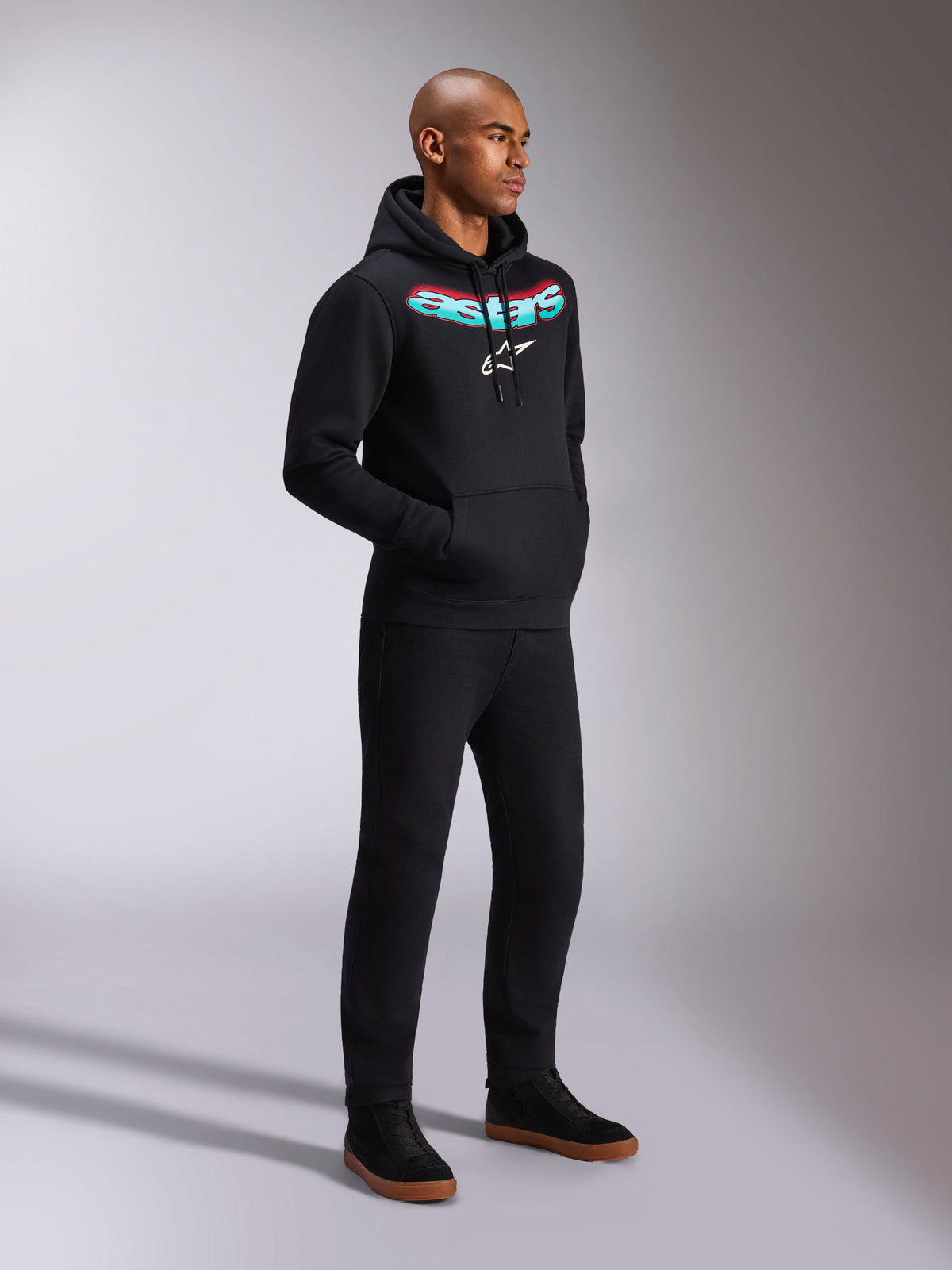 Alpinestars Ellipsoid Hoodie, camisola com capuz em fleece preto, com a palavra Astars curvada em gradiente verde-azulado e vermelho e logótipo de estrela branco, usada por um modelo com calças pretas, streetwear casual inspirado no desporto motorizado