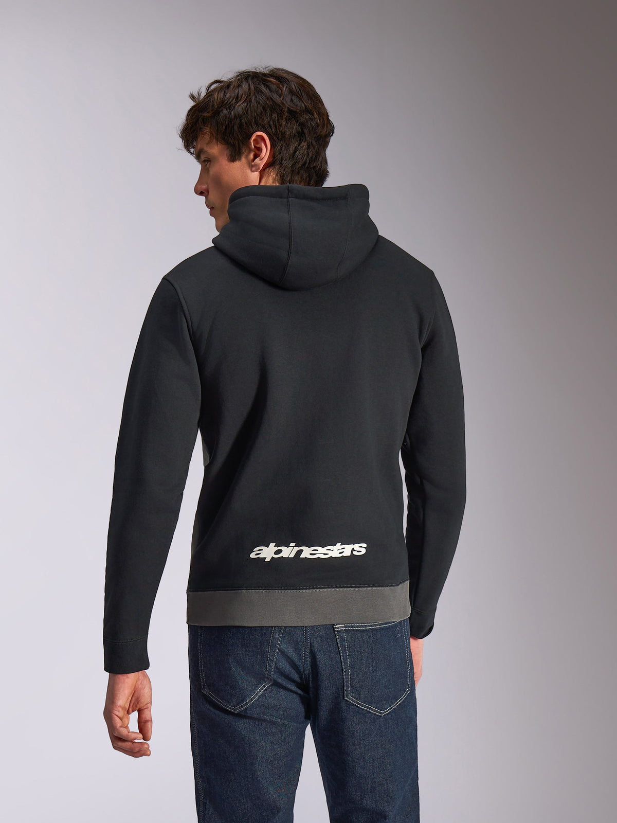 Alpinestars Parallel Hoodie, sweatshirt casual de sportswear com capuz, preto e gunmetal, vista traseira com logótipo Alpinestars branco na parte inferior das costas, design em blocos de cor com bainha cinzenta, usada com calças de ganga escuras
