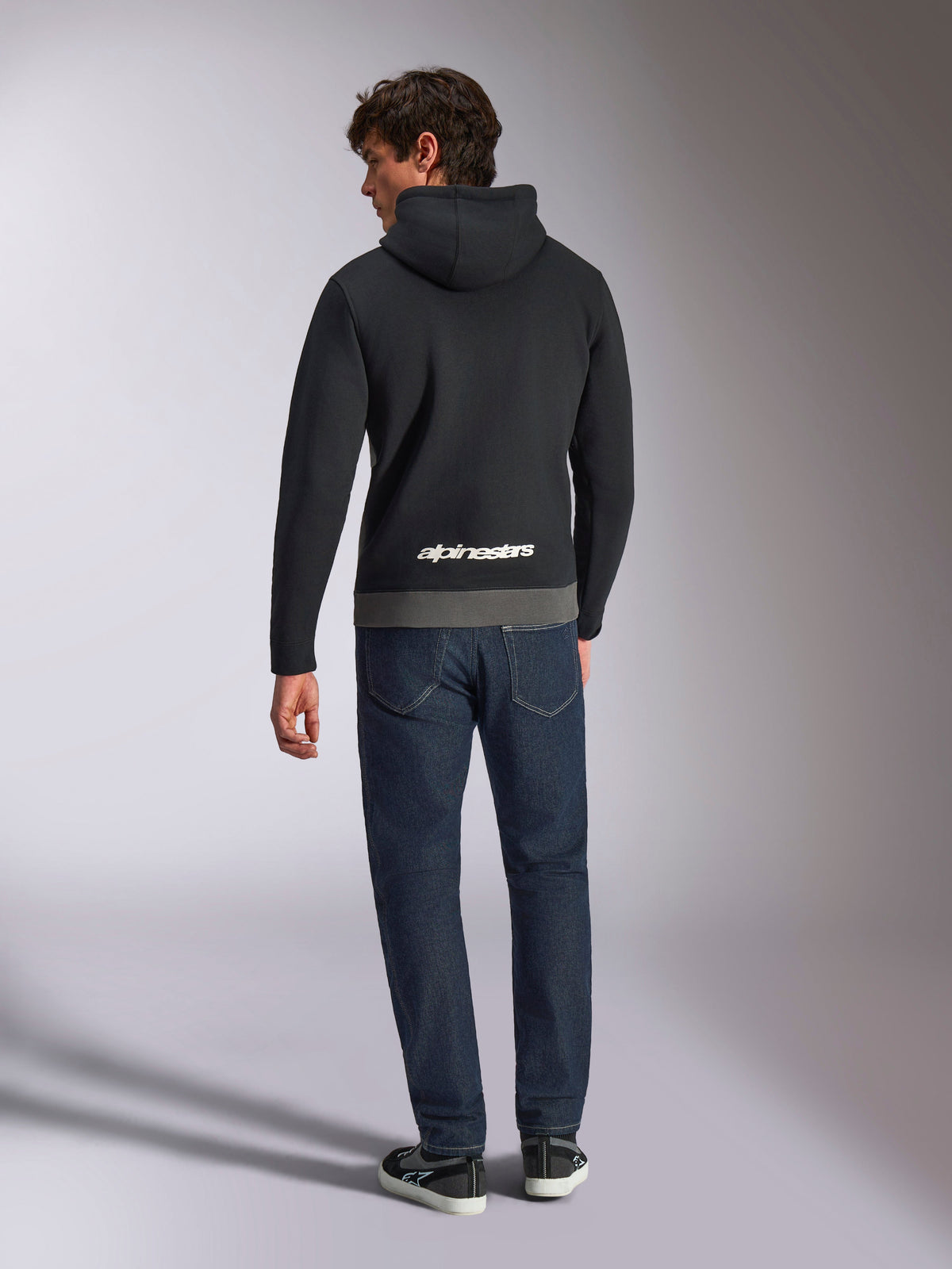 Alpinestars Parallel Hoodie, vista traseira de um homem a usar uma camisola com capuz em fleece com blocos de cor preto e cinzento gunmetal, logótipo da palavra Alpinestars em branco na parte inferior das costas, combinada com calças de ganga azul escuras, streetwear casual inspirado no desporto motorizado