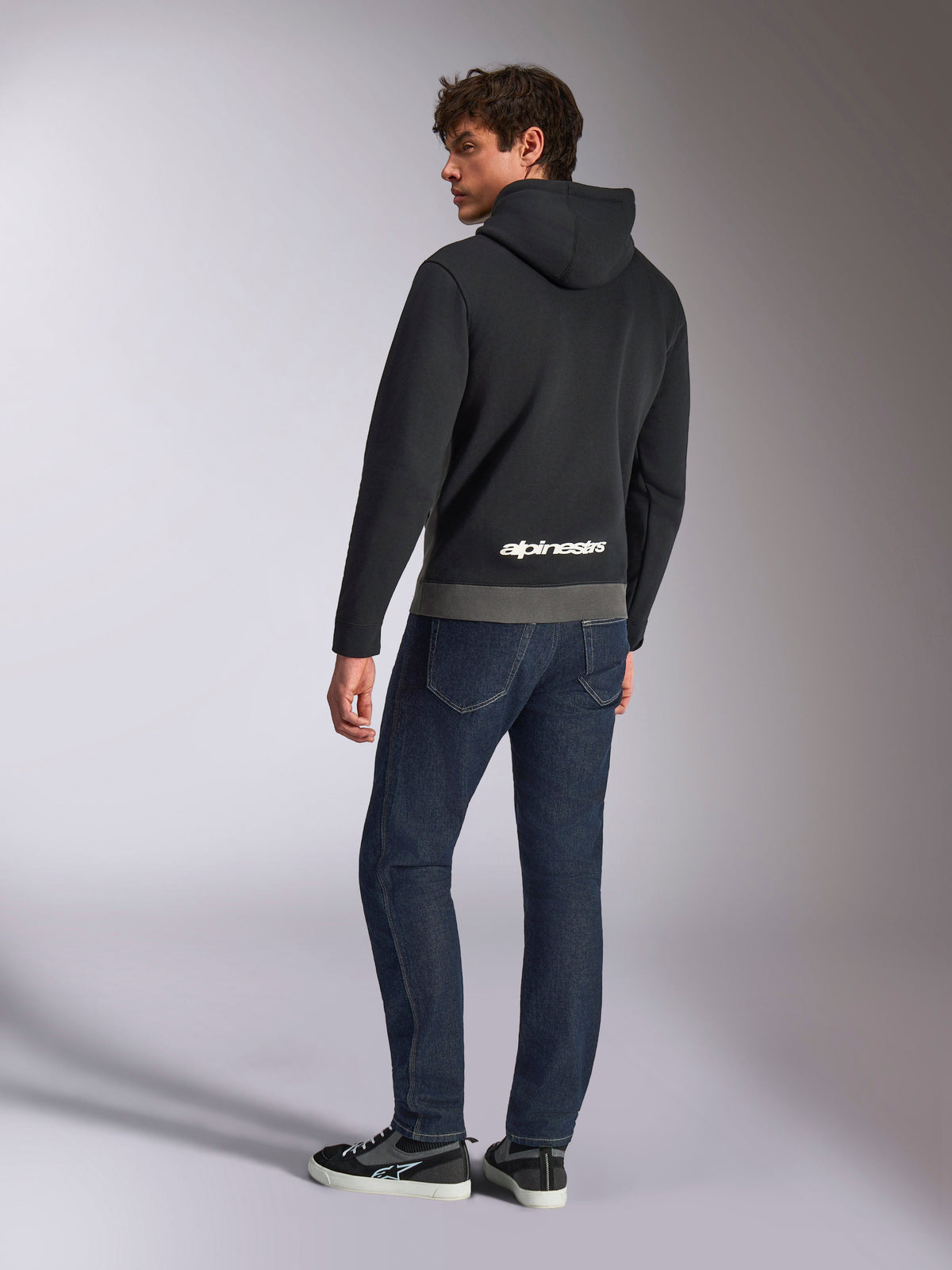 Alpinestars Parallel Hoodie, vista traseira do modelo a usar a sweatshirt preta e cinzento gunmetal, com logótipo Alpinestars branco na parte inferior das costas, combinado com calças de ganga azul escuro, streetwear casual inspirado no desporto motorizado