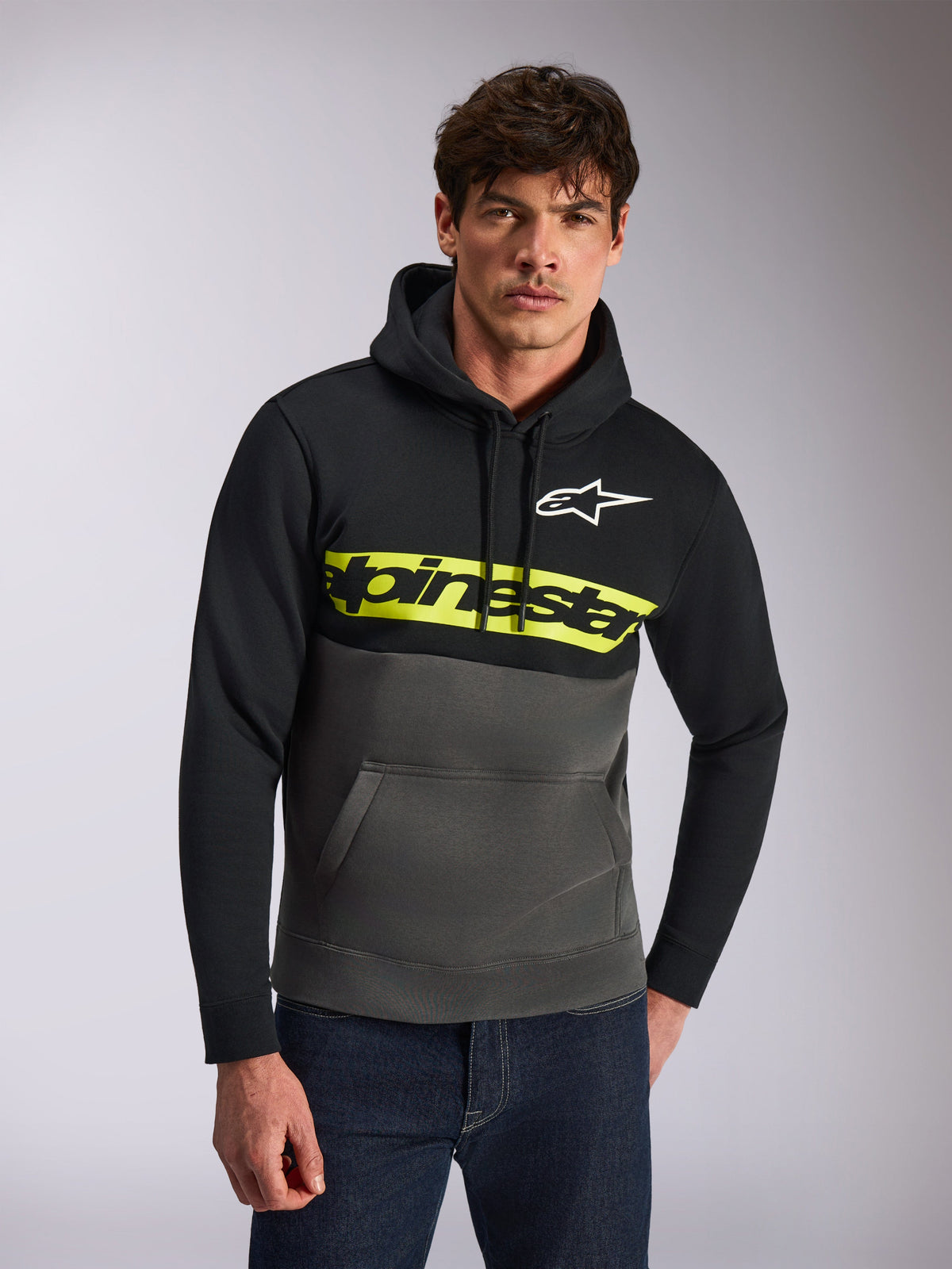 Alpinestars Parallel Hoodie, camisola com capuz de streetwear casual, design em blocos de cor preto e cinzento gunmetal, faixa amarela vibrante no peito com logótipo Alpinestars, logótipo Astars branco, vestido por um modelo com calças de ganga azuis, design moderno inspirado no desporto motorizado