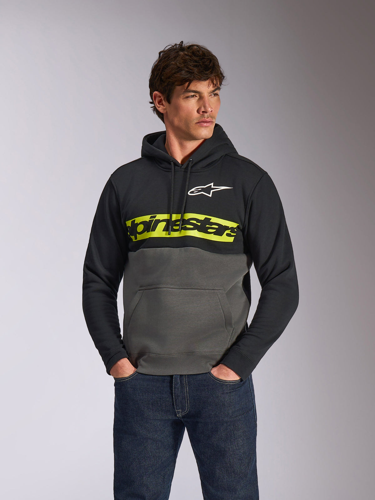 Alpinestars Parallel Hoodie, sweatshirt em fleece casual com design de blocos de cor, preto e gunmetal com uma barra amarela vibrante no peito, logótipo Alpinestars e logótipo da estrela branco, vestido por um modelo masculino com calças de ganga azul-escuro