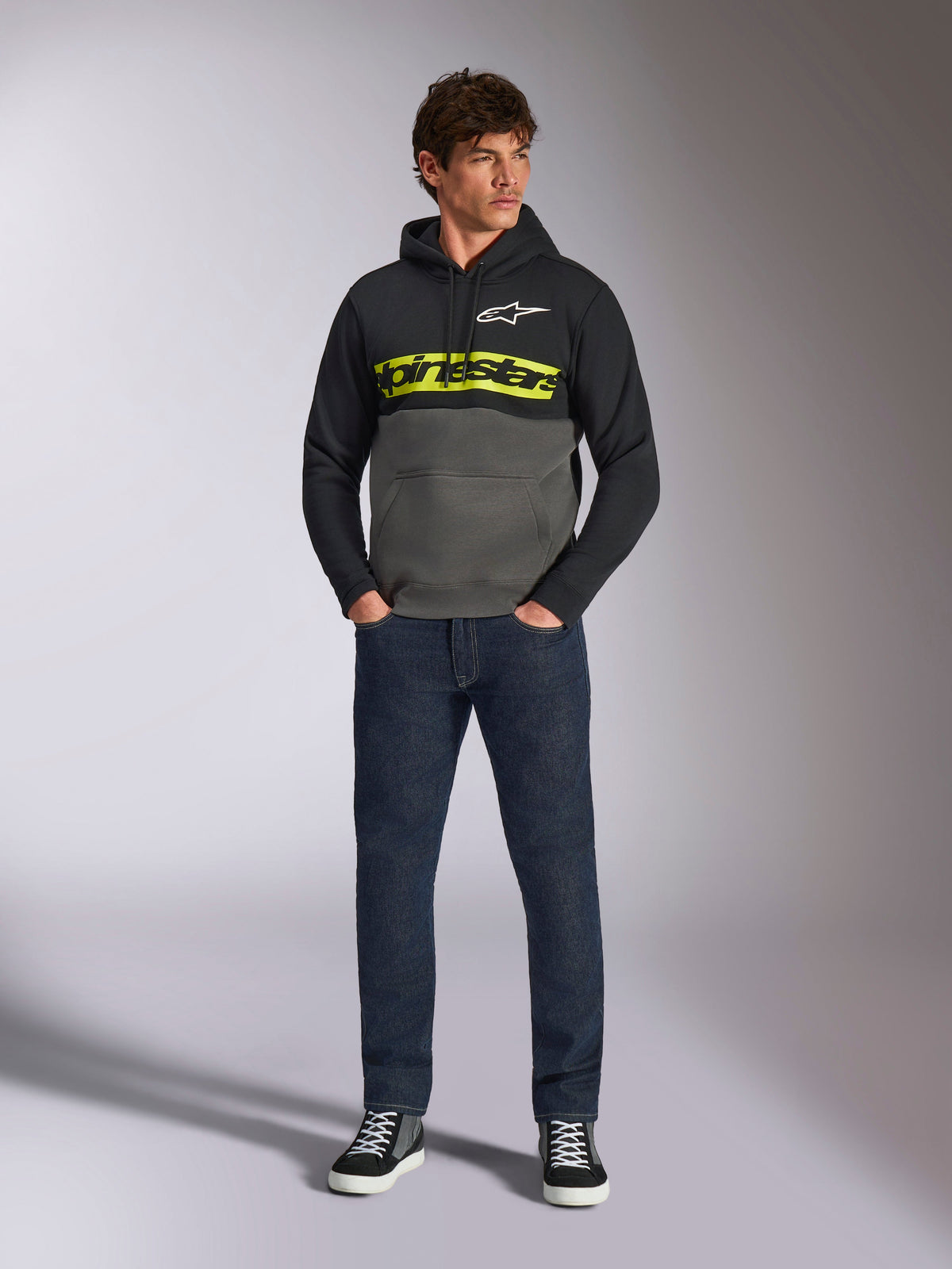 Alpinestars Parallel Hoodie, camisola de capuz com blocos de cor, preto e cinzento Gunmetal, logótipo Alpinestars em amarelo vibrante e logótipo de estrela branco no peito, vestido por um modelo masculino com calças de ganga azul-escuro e sapatilhas pretas, streetwear casual de inspiração motorsport