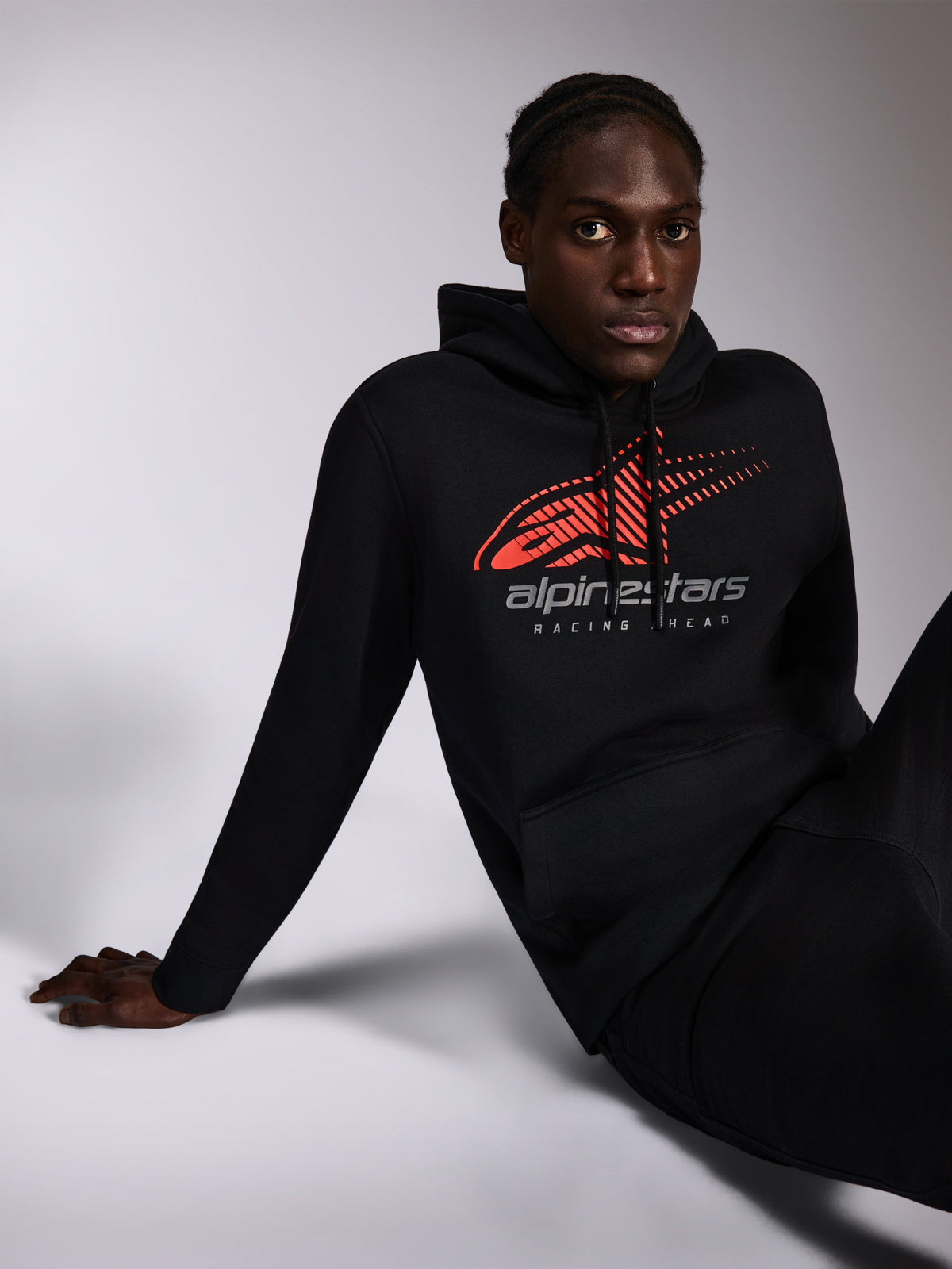 Alpinestars Pulse Hoodie, camisola com capuz preta, estilo casual streetwear usado por um modelo, com logótipo Astars frontal vermelho com efeito speed line fade e texto 'alpinestars RACING AHEAD' no peito, capuz com cordão, mangas compridas, design moderno inspirado no desporto motorizado