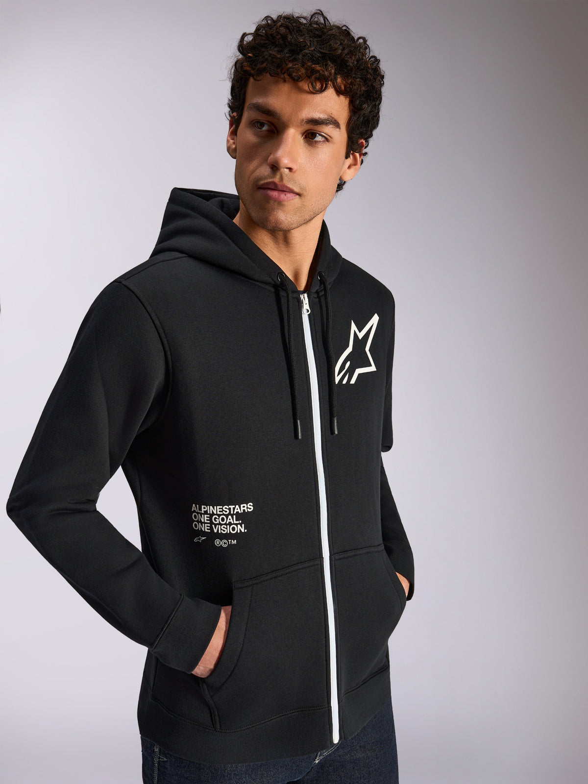 Alpinestars Transverse Hoodie, camisola desportiva casual, preta com detalhe de fecho branco, capuz com cordão, logótipo de estrela Alpinestars no peito, grafismo 'One Goal One Vision', usada por modelo masculino com cabelo escuro encaracolado