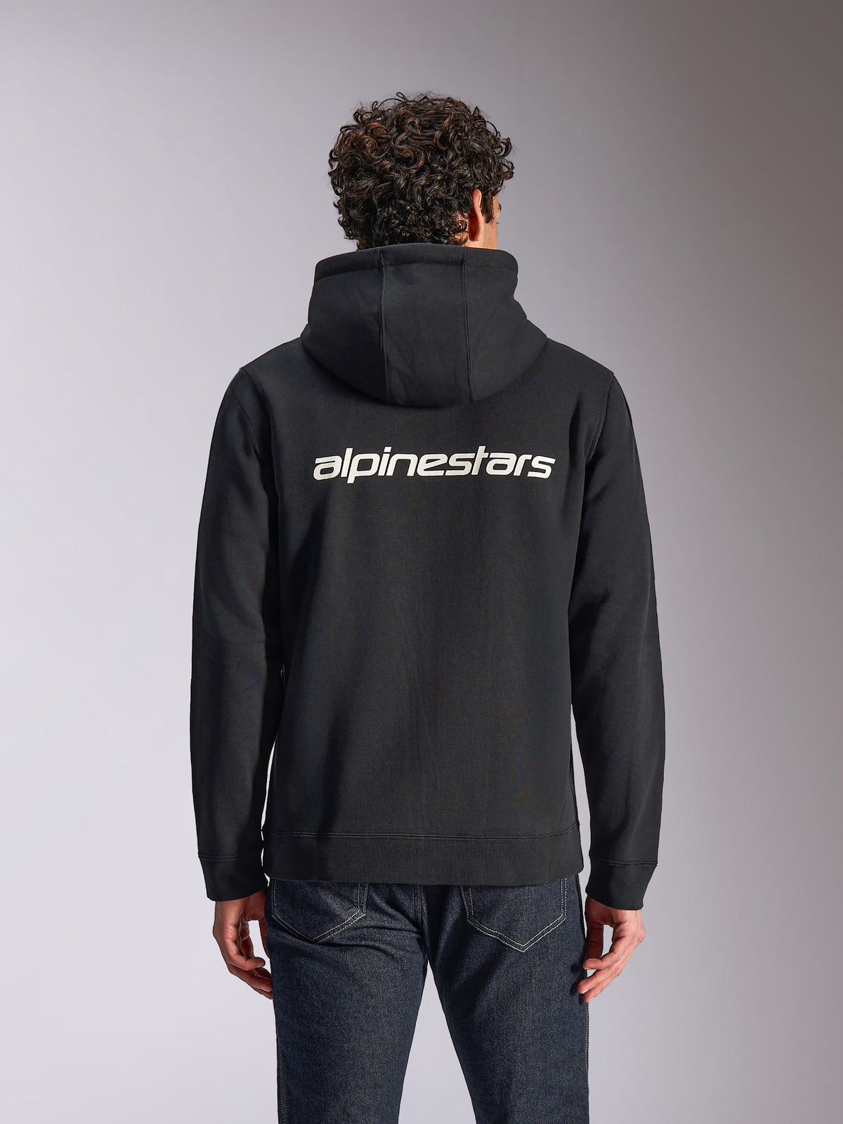Alpinestars Transverse Hoodie, estilo casual streetwear, preta com logótipo Alpinestars branco nas costas, camisola de fleece com capuz e fecho frontal, mangas compridas, usada por um modelo com calças de ganga escuras, design moderno inspirado no desporto motorizado