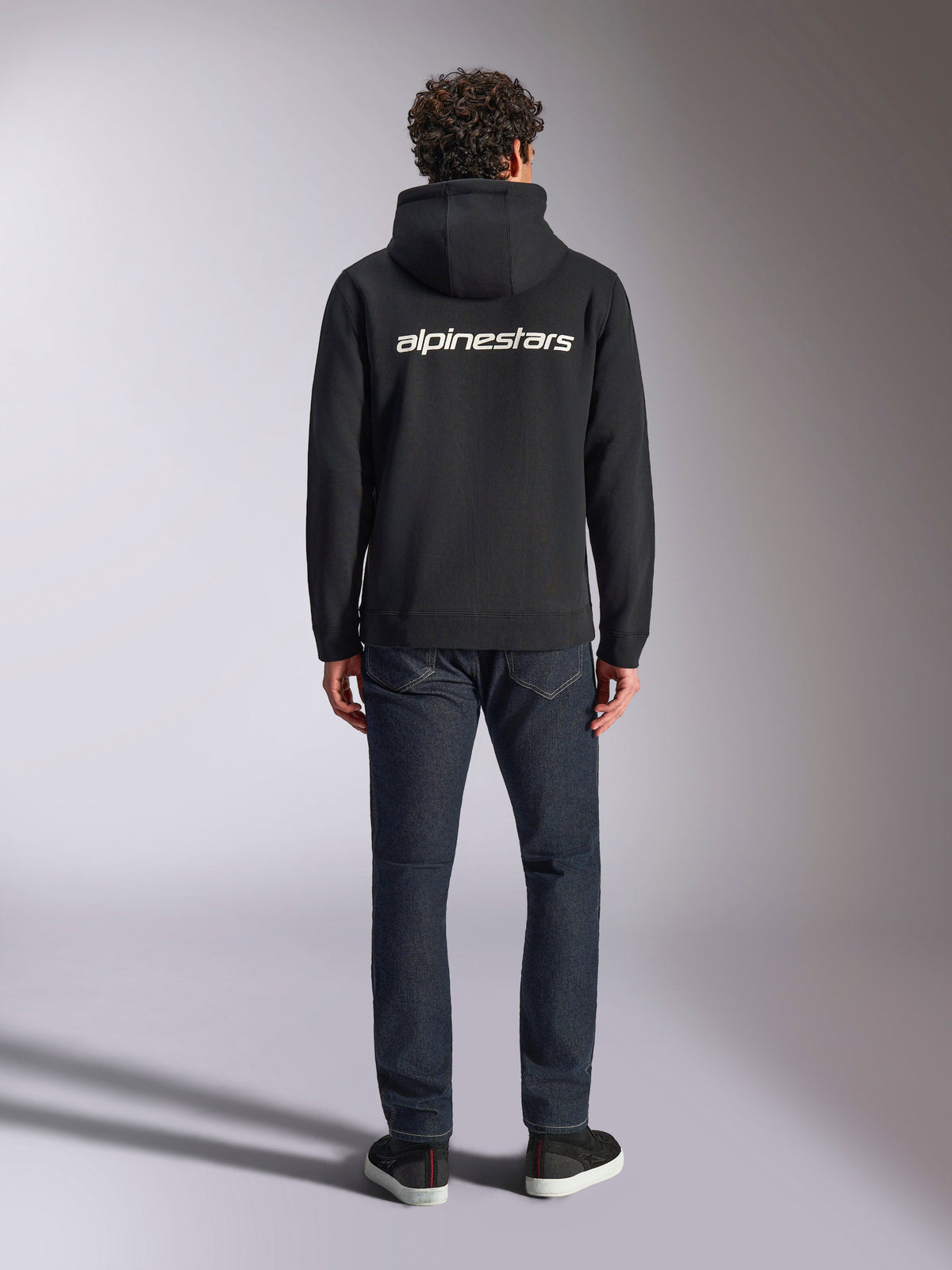 Alpinestars Transverse Hoodie, streetwear casual de desportos motorizados, preta com logótipo Alpinestars branco nas costas, mangas compridas, bainha e punhos canelados, vista de costas com calças de ganga escuras