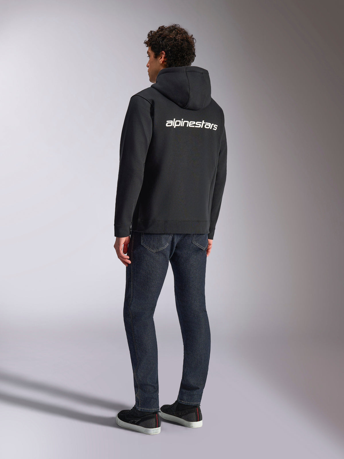 Alpinestars Transverse Hoodie, fleece preto com fecho frontal, vista traseira com logótipo Alpinestars em branco nos ombros, usado com calças de ganga azul-escuras e ténis pretos, streetwear casual inspirado no desporto motorizado