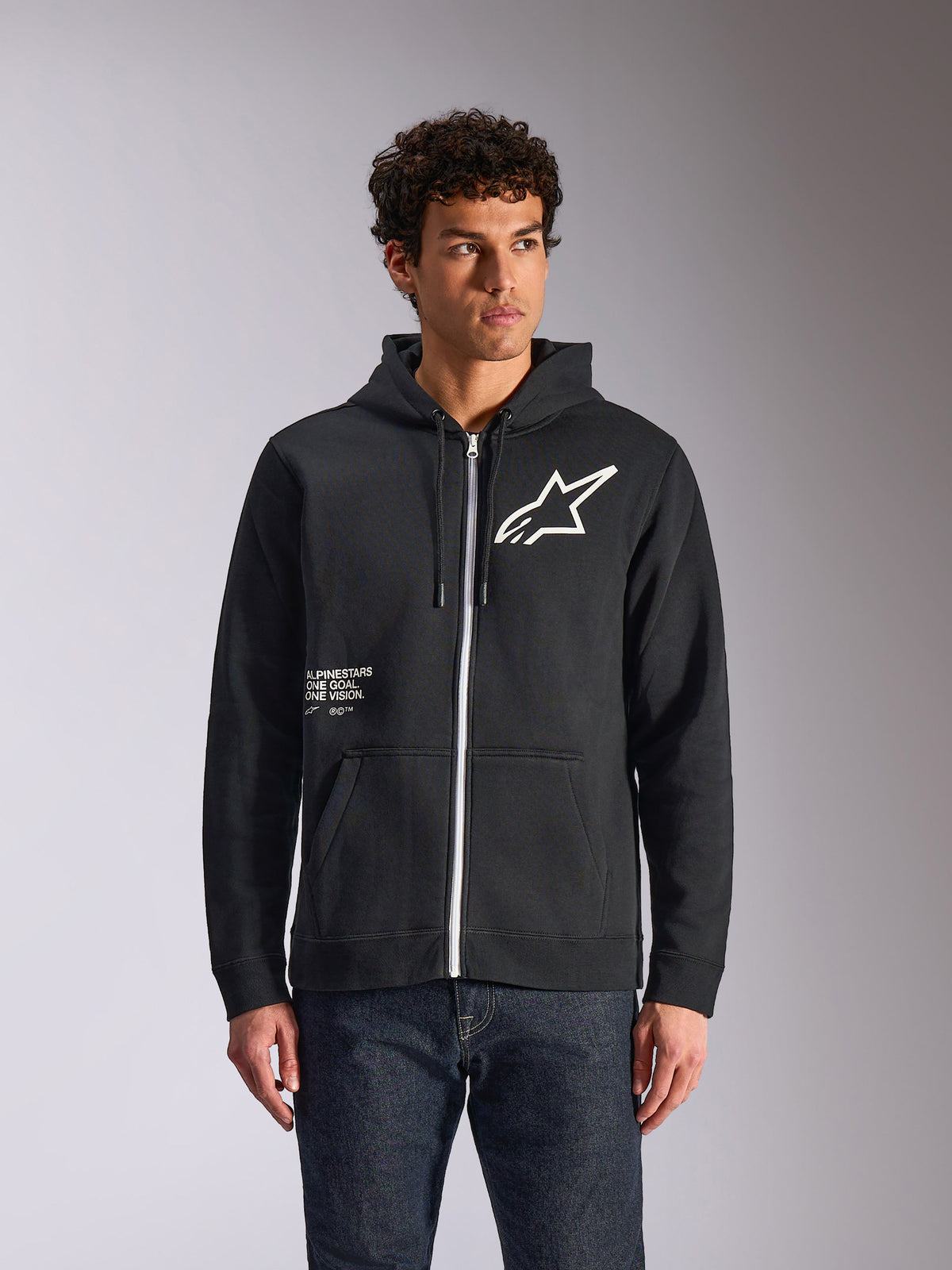 Alpinestars Transverse Hoodie, sweatshirt preta com capuz e fecho frontal em fleece, com o logótipo Astars branco e o grafismo One Goal na frente, usada por um modelo com calças de ganga escuras, streetwear casual inspirado no desporto motorizado