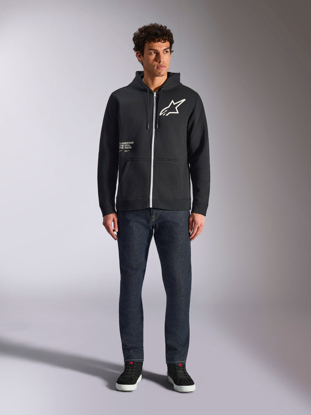 Alpinestars Transverse Hoodie, camisola casual de fleece com fecho frontal, preta, logótipo Astars branco no peito e impressão 'One Goal' na frente, capuz com cordão, combinada com calças de ganga escuras e sapatilhas pretas, lifestyle motorsport streetwear